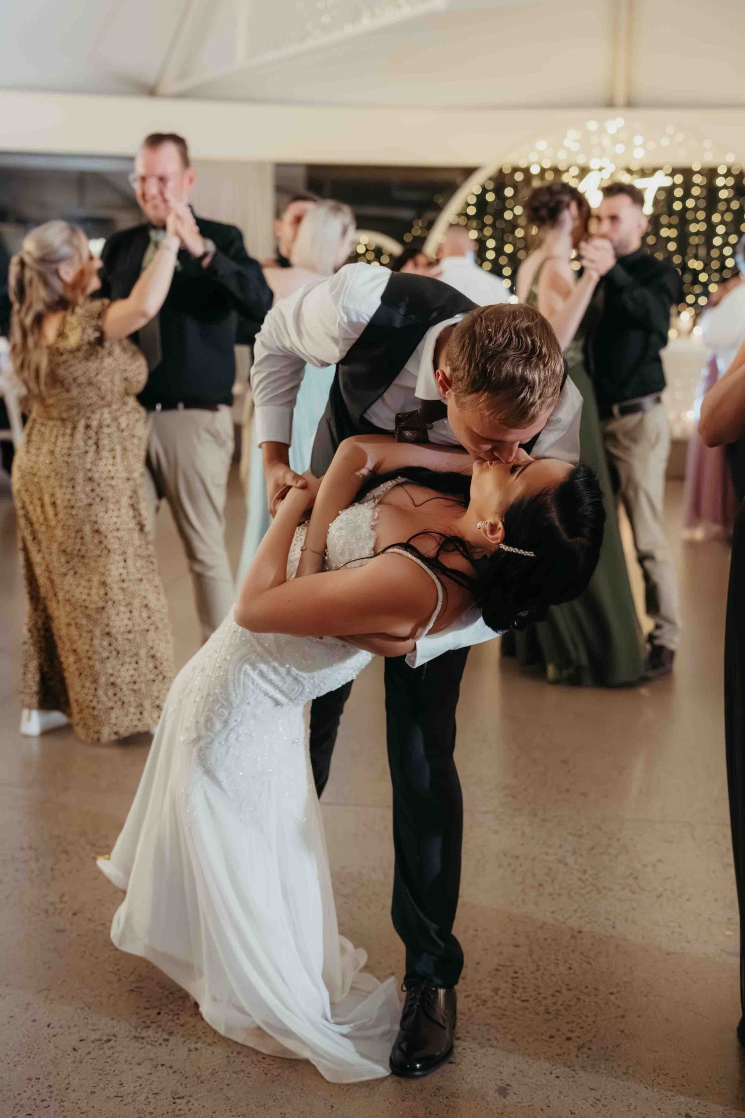 Bride and groom share a romantic dip kiss on the wedding dance floor surrounded by guests celebrating.