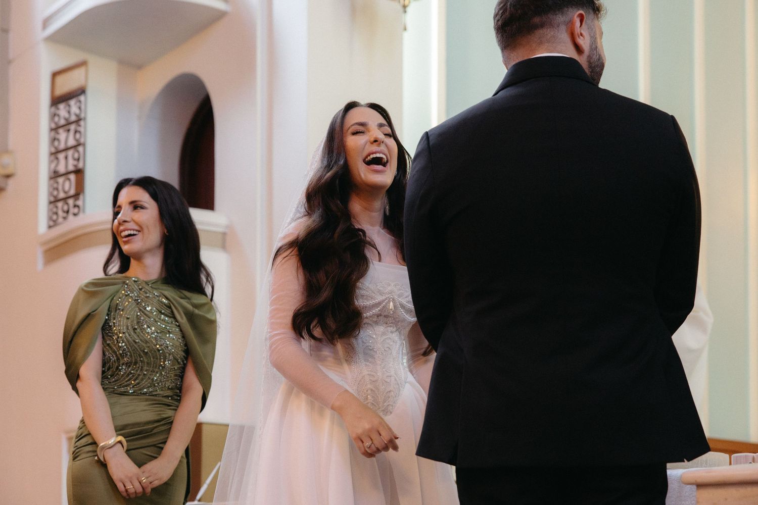 Bride laughing joyfully at wedding ceremony altar in white gown, bridesmaid in green dress smiling nearby.
