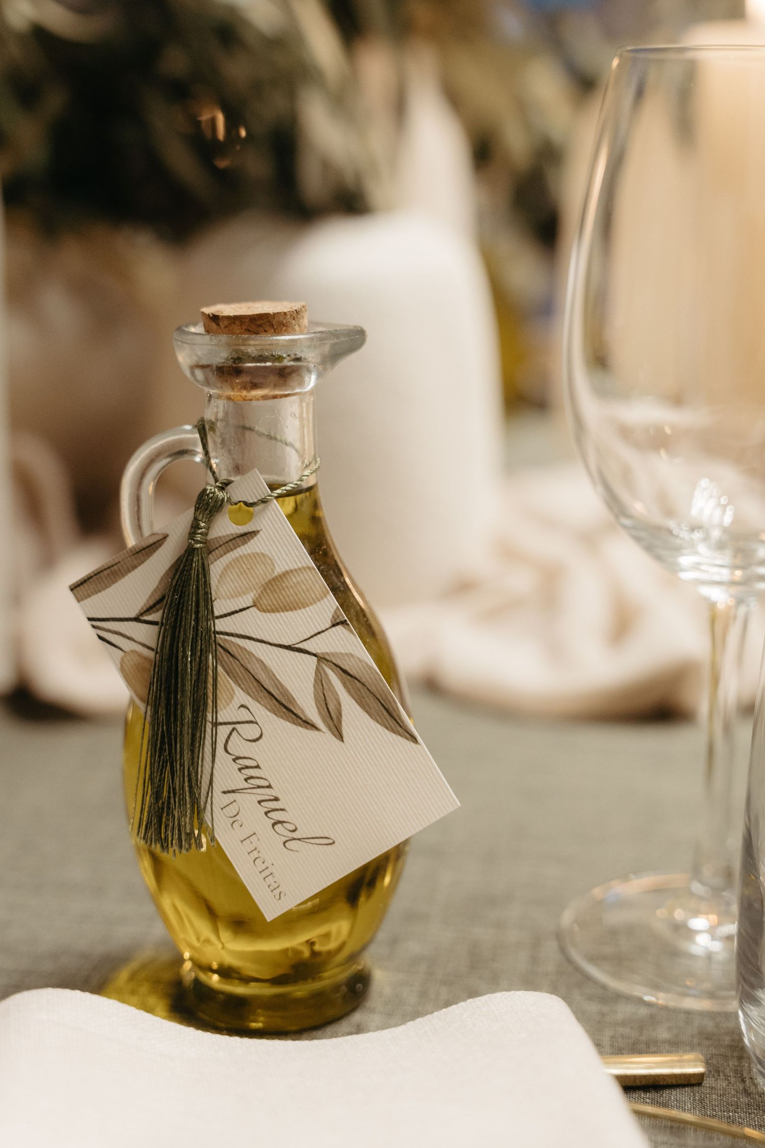 Elegant olive oil bottle wedding favor with tassel and botanical tag on a gray linen tablescape.