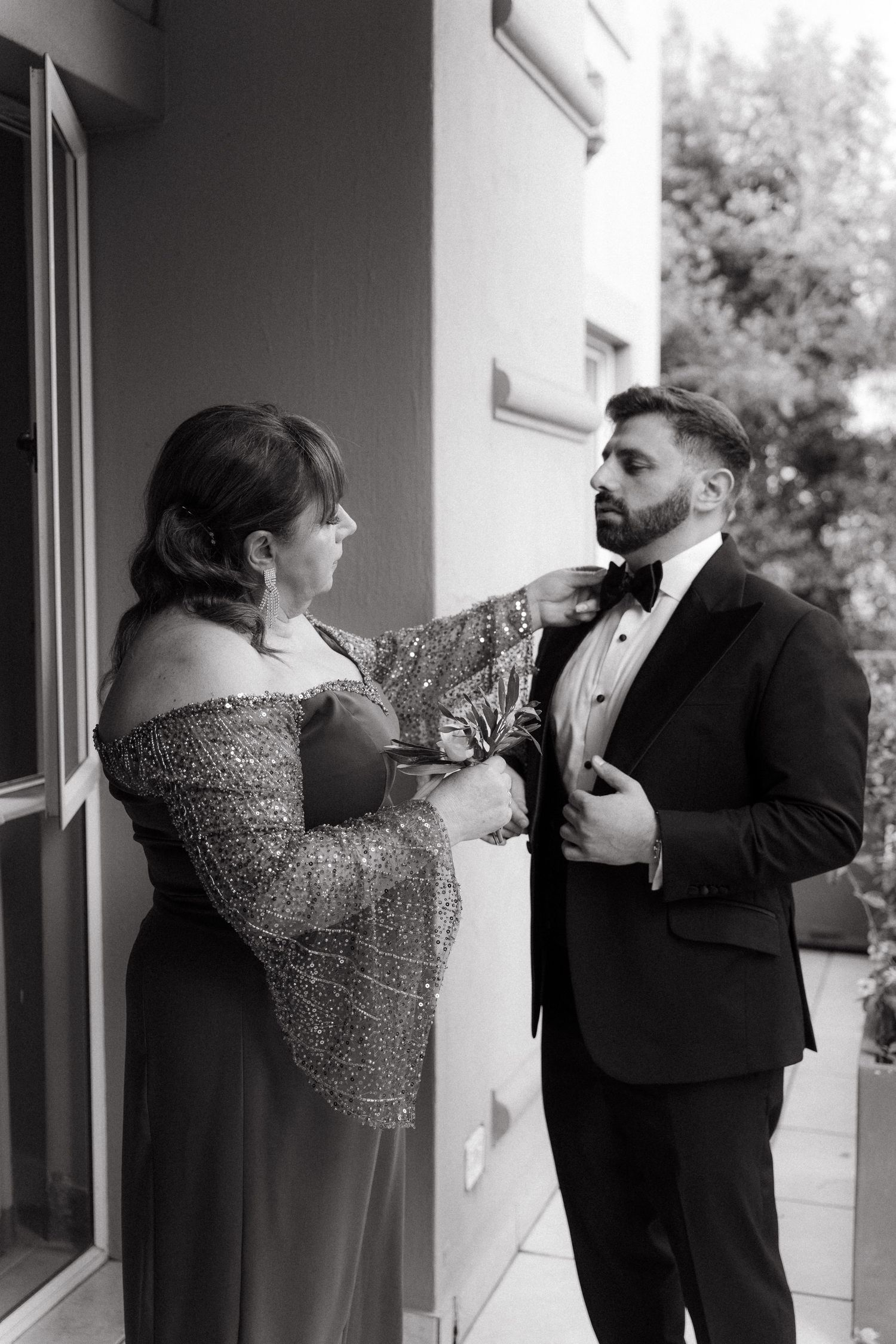 Black and white photo of a woman in sequin gown adjusting a groom's bow tie before his Johannesburg wedding ceremony.