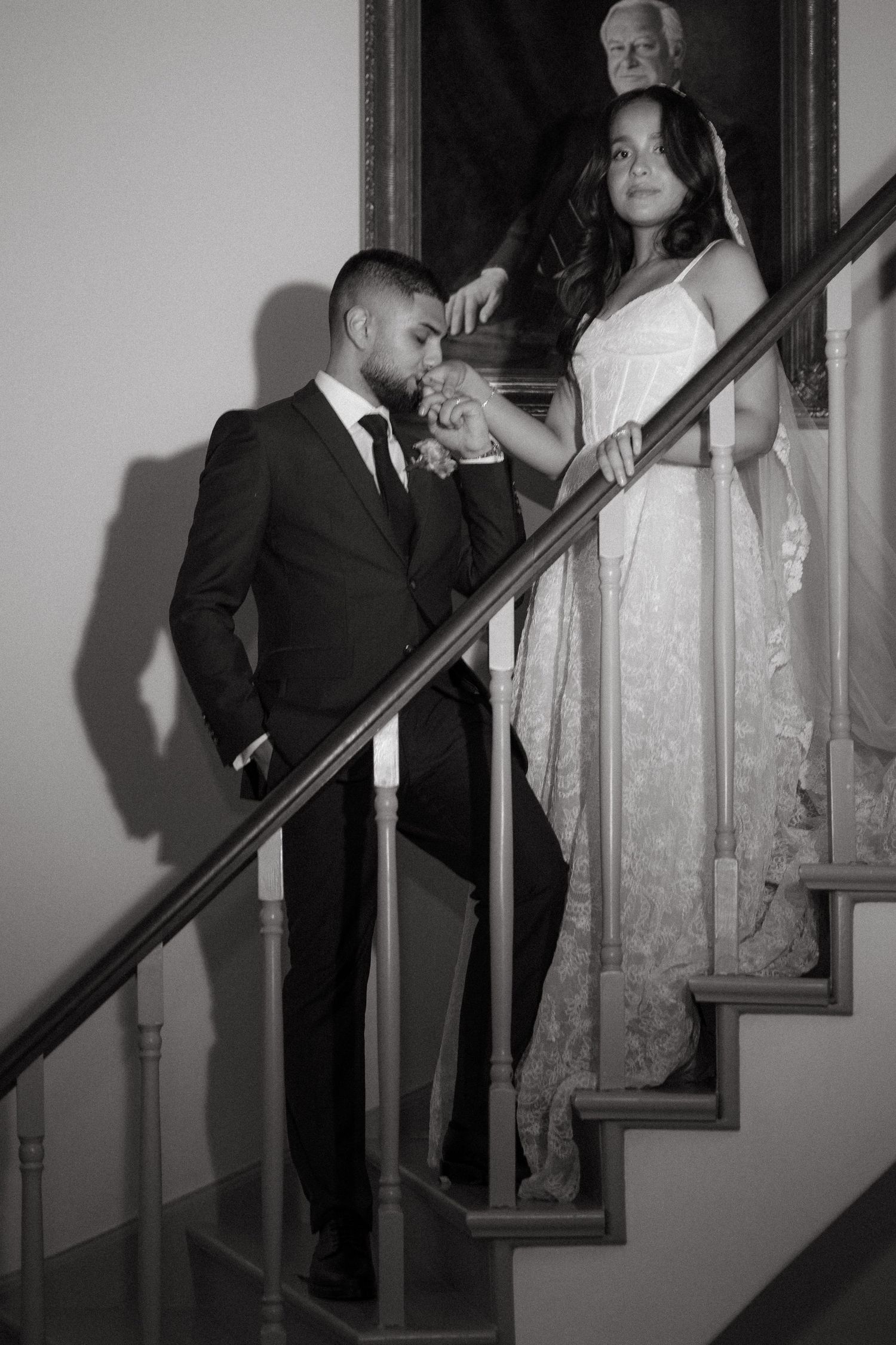 Black and white wedding photo of groom kissing bride's hand on elegant staircase with portrait painting backdrop.