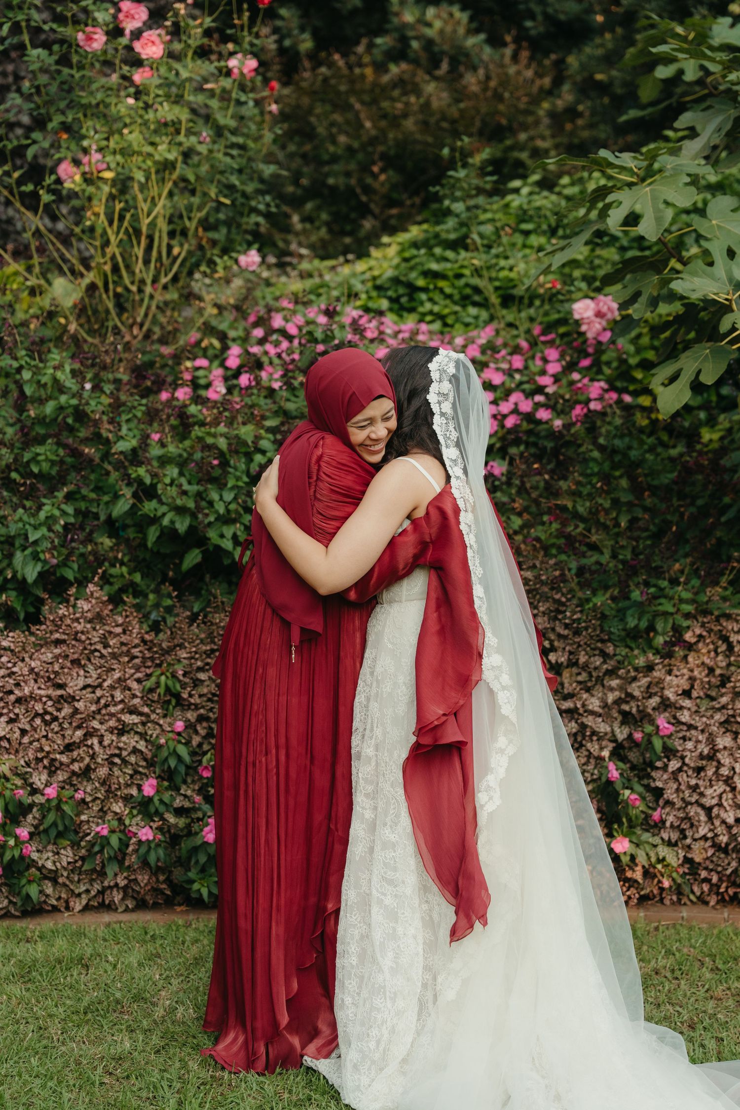 Bride in white lace gown and veil embracing a woman in burgundy hijab and dress, surrounded by pink roses.