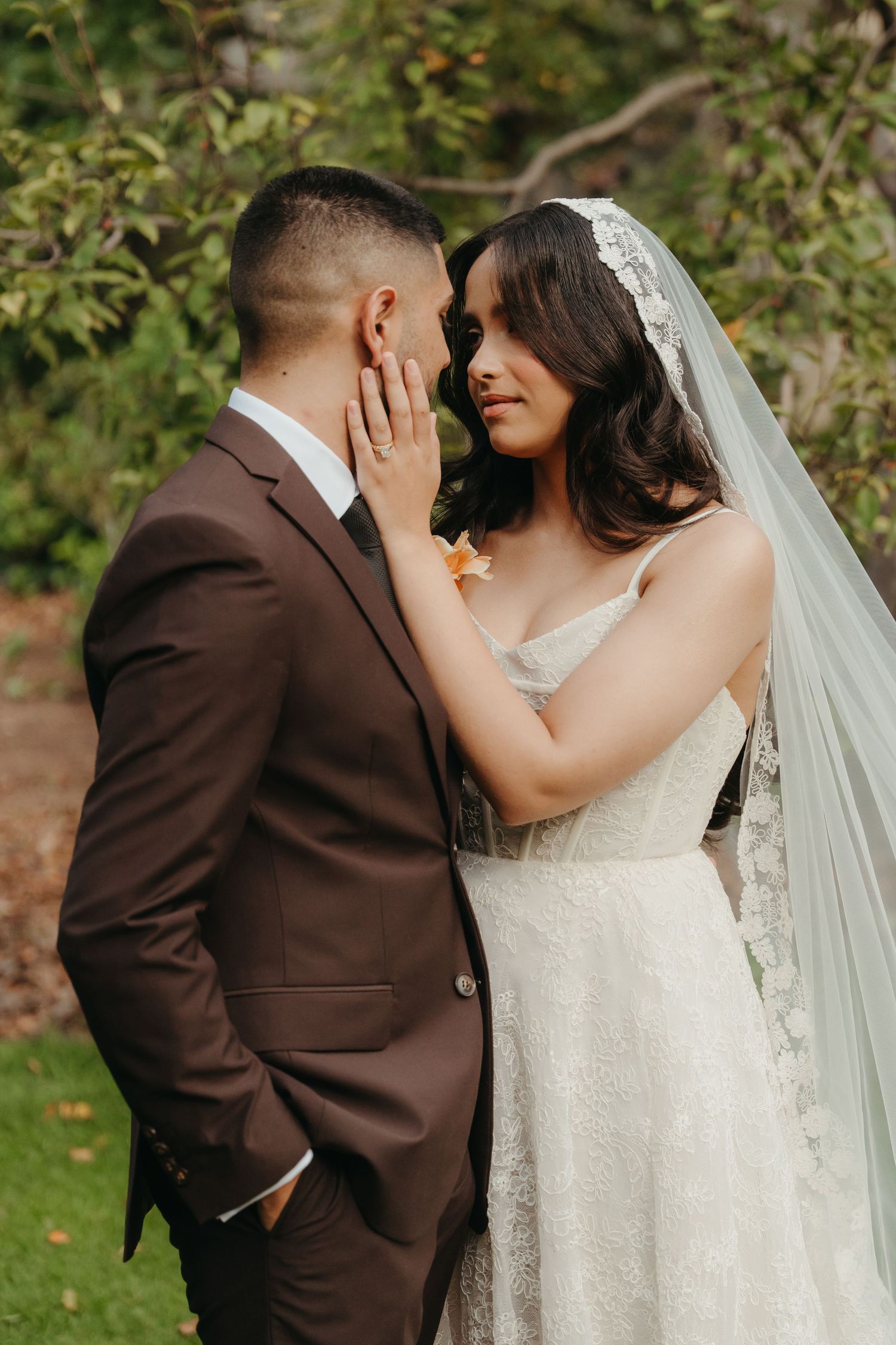 A bride in a lace gown and veil tenderly touches the groom's face as they share an intimate moment outdoors.