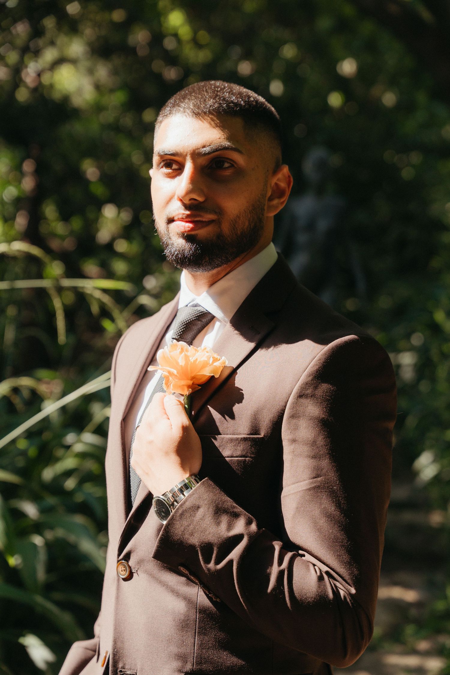 Groom in brown suit with orange boutonniere posing outdoors in garden setting.