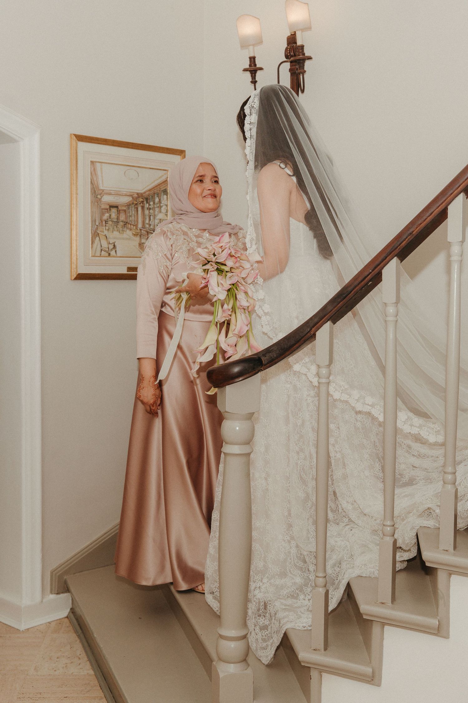 Bride in lace gown and veil holds floral bouquet on elegant staircase with bridesmaid in rose satin hijab dress.