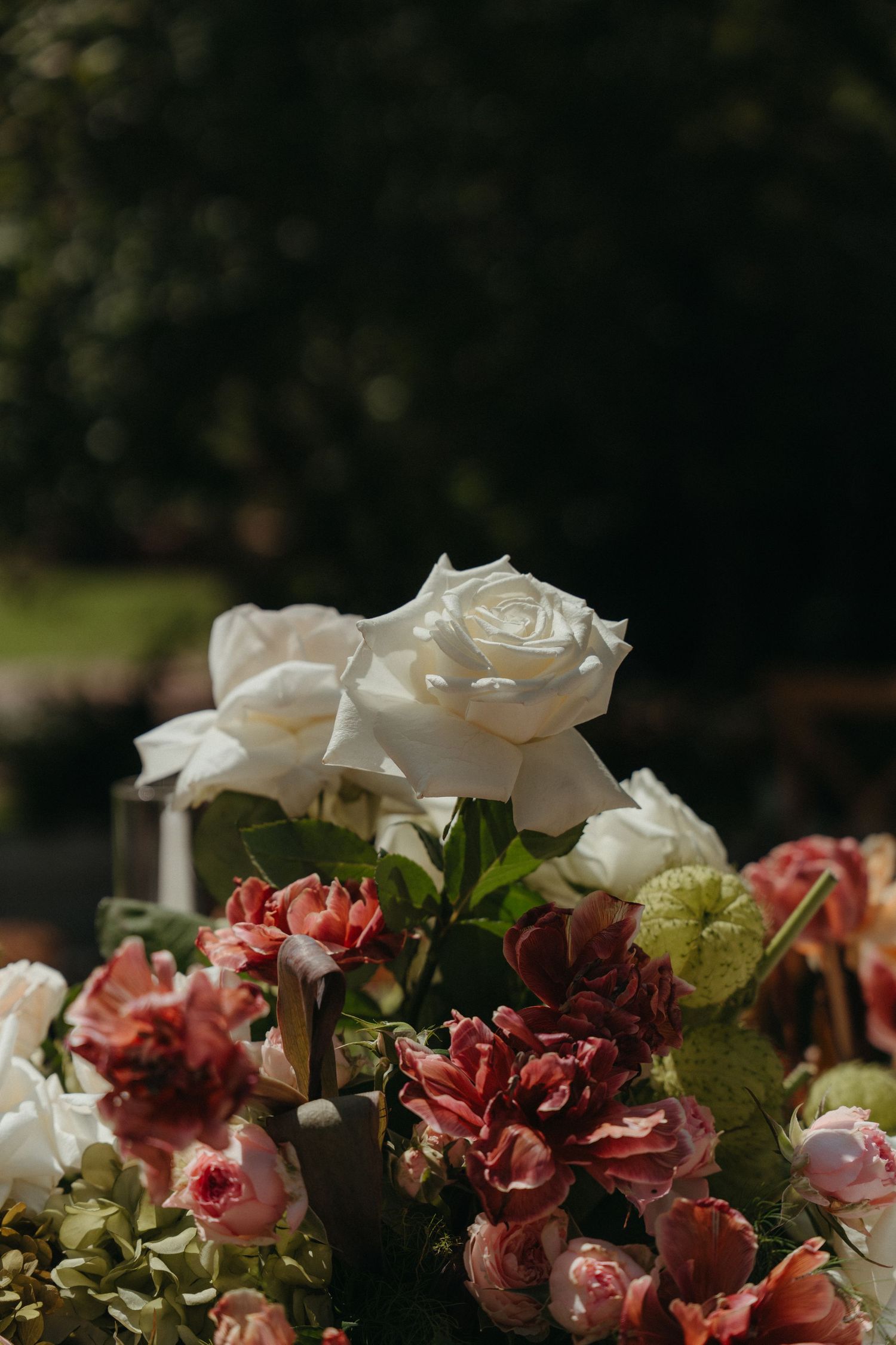 White roses stand above pink and red blooms in an elegant outdoor floral arrangement with dark foliage background.