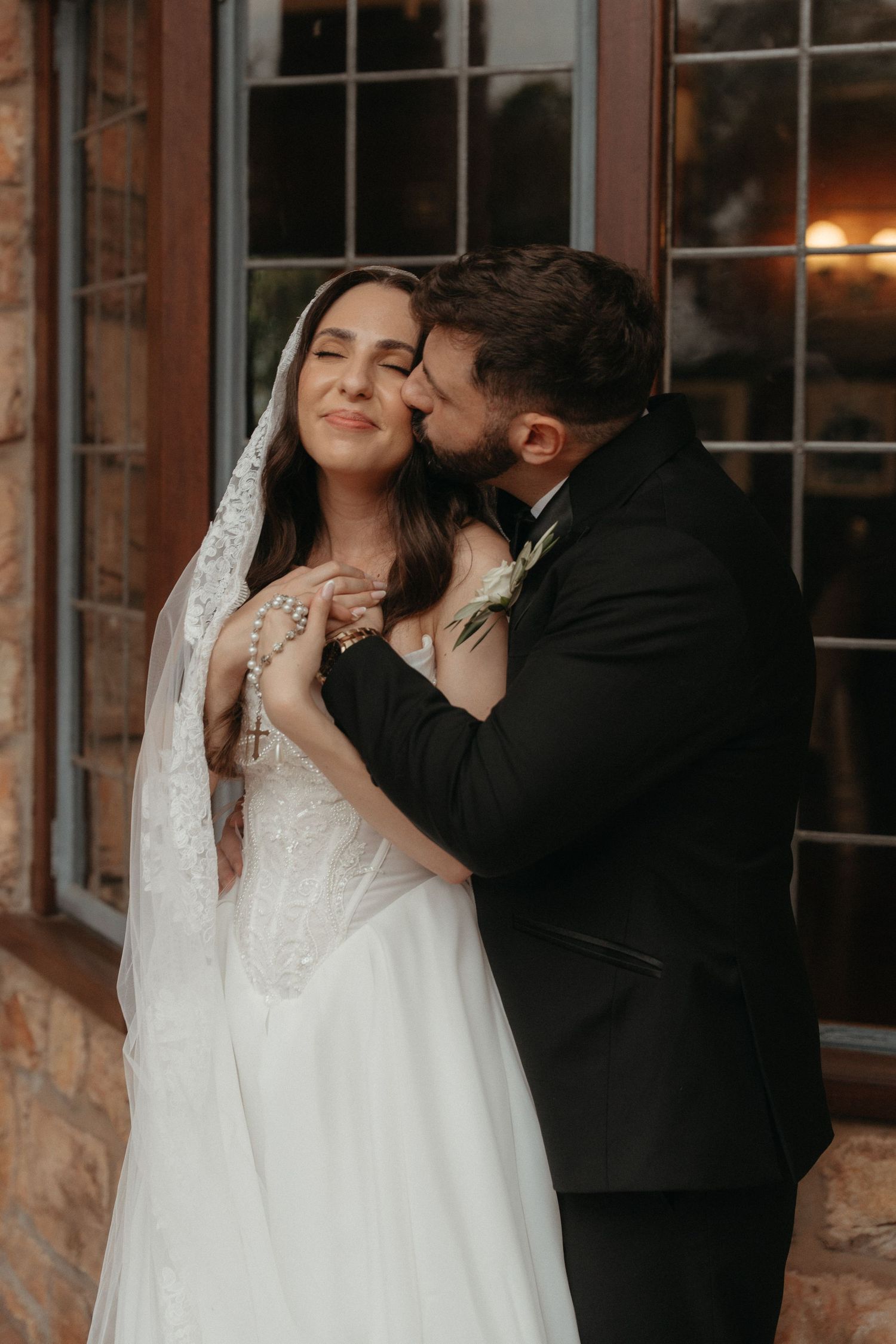 Groom in black tuxedo kissing bride in white lace wedding dress and veil outside Glenshiel in Johannesburg