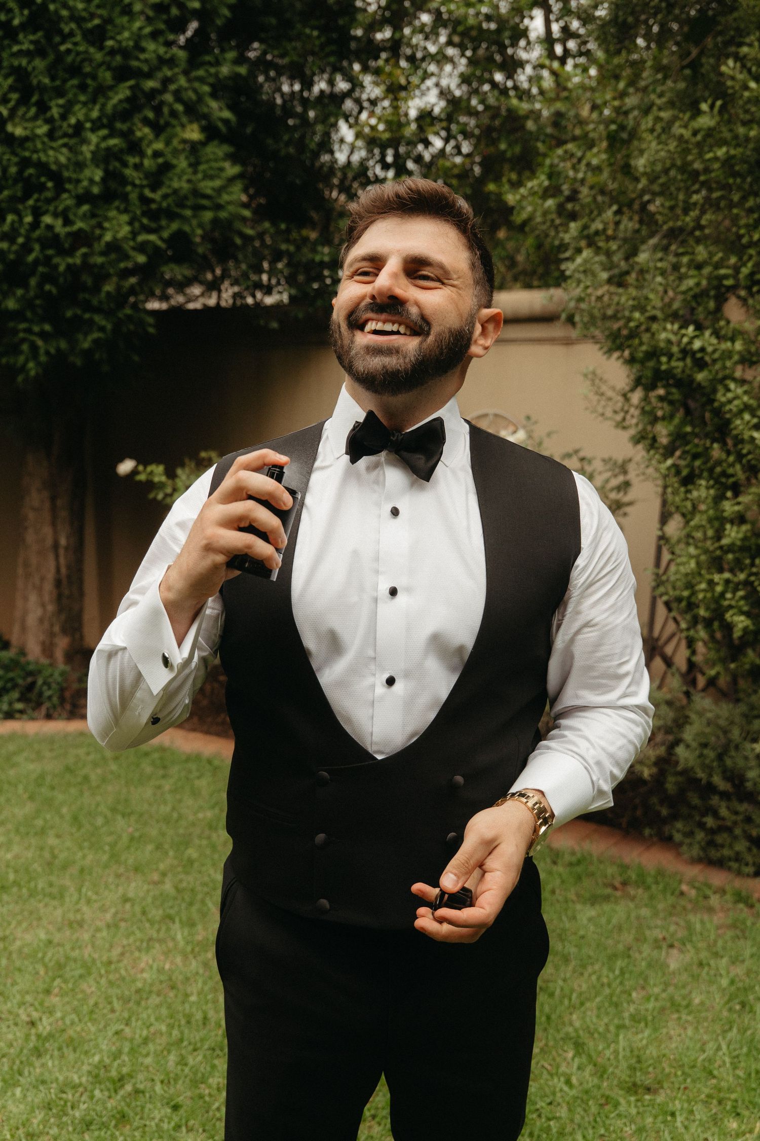 Smiling groom in tuxedo vest and bow tie spraying cologne outdoors in a garden setting, Johannesburg Wedding Photographer