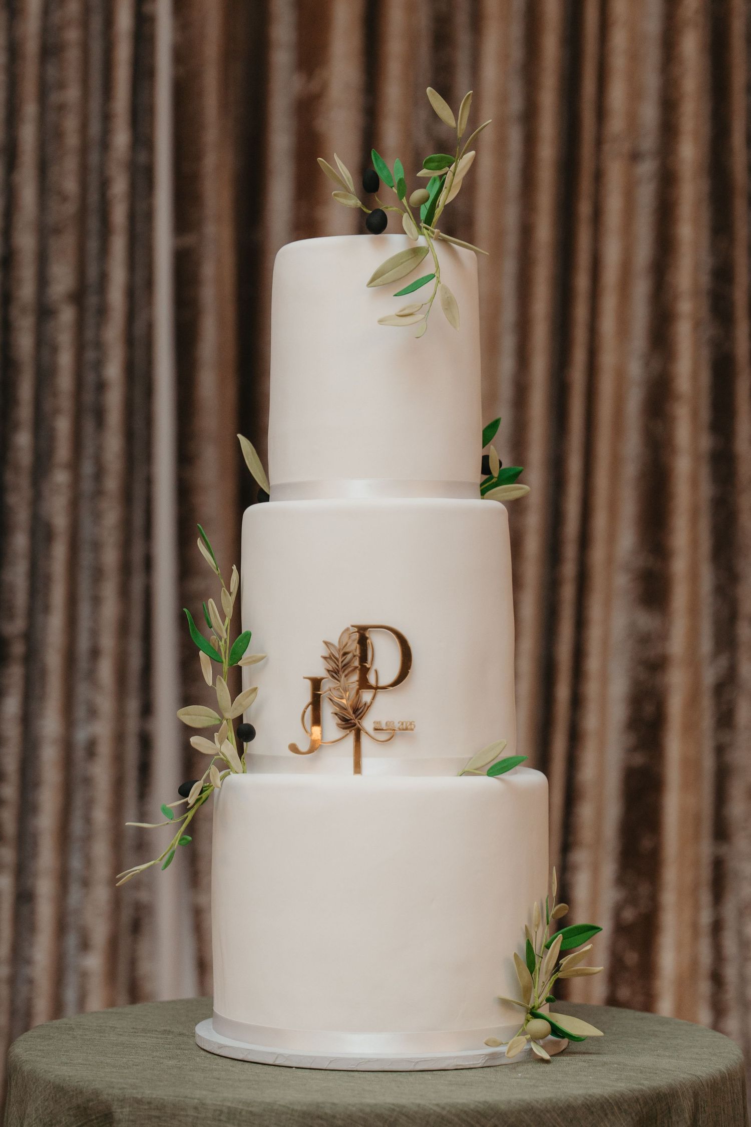 Elegant three-tier white wedding cake with gold monogram and olive branch accents on a green stand.