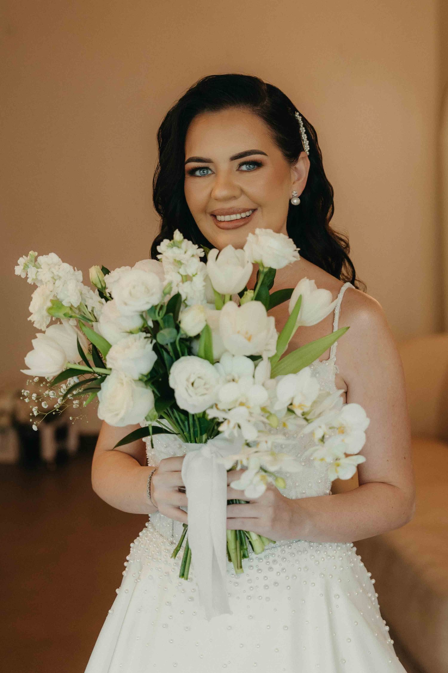 Smiling bride in white gown holding elegant bouquet of white tulips and roses, with pearl hair accessories.