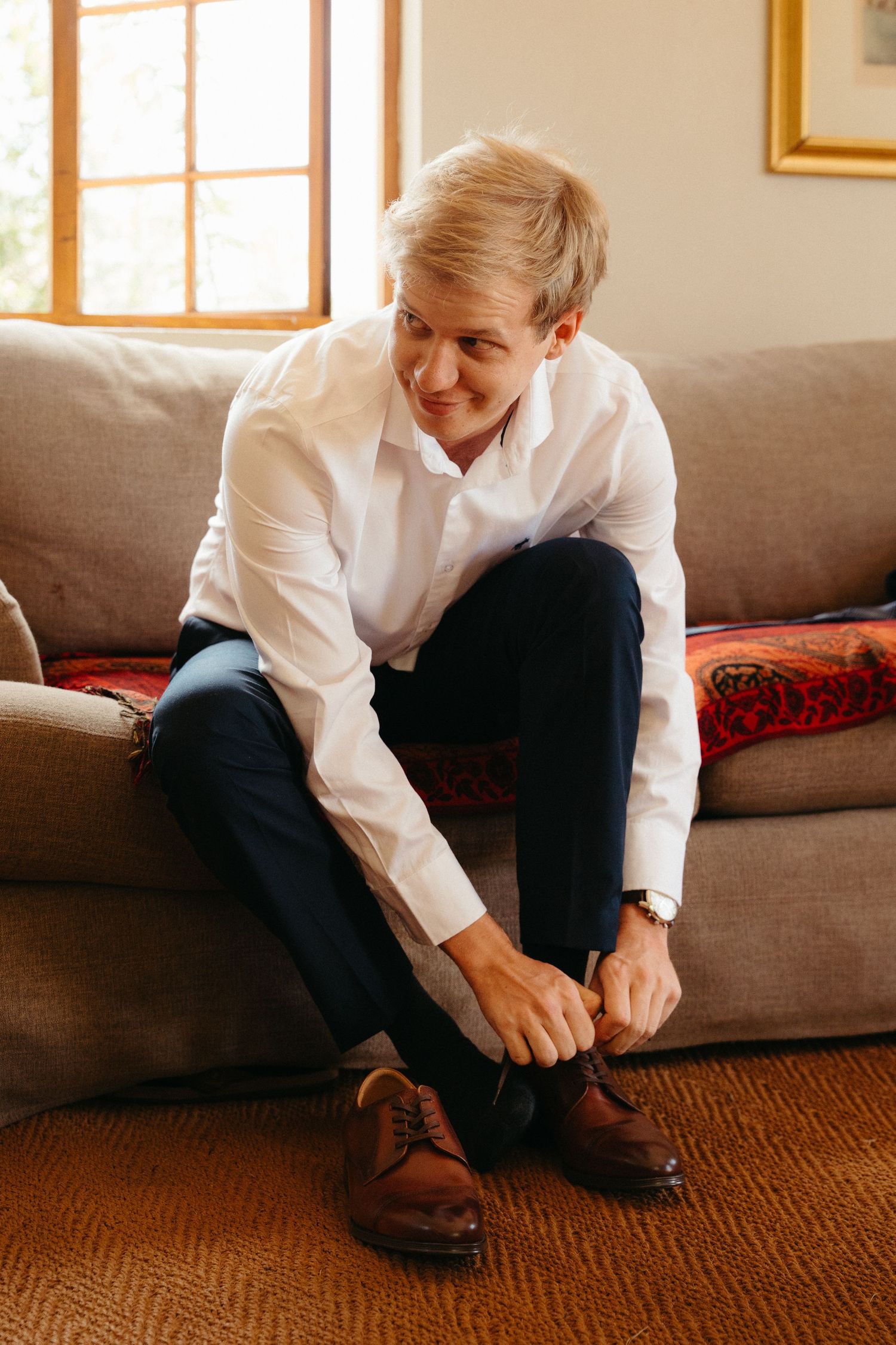 Someone in a white shirt sits on a brown couch while tying their brown leather shoes.