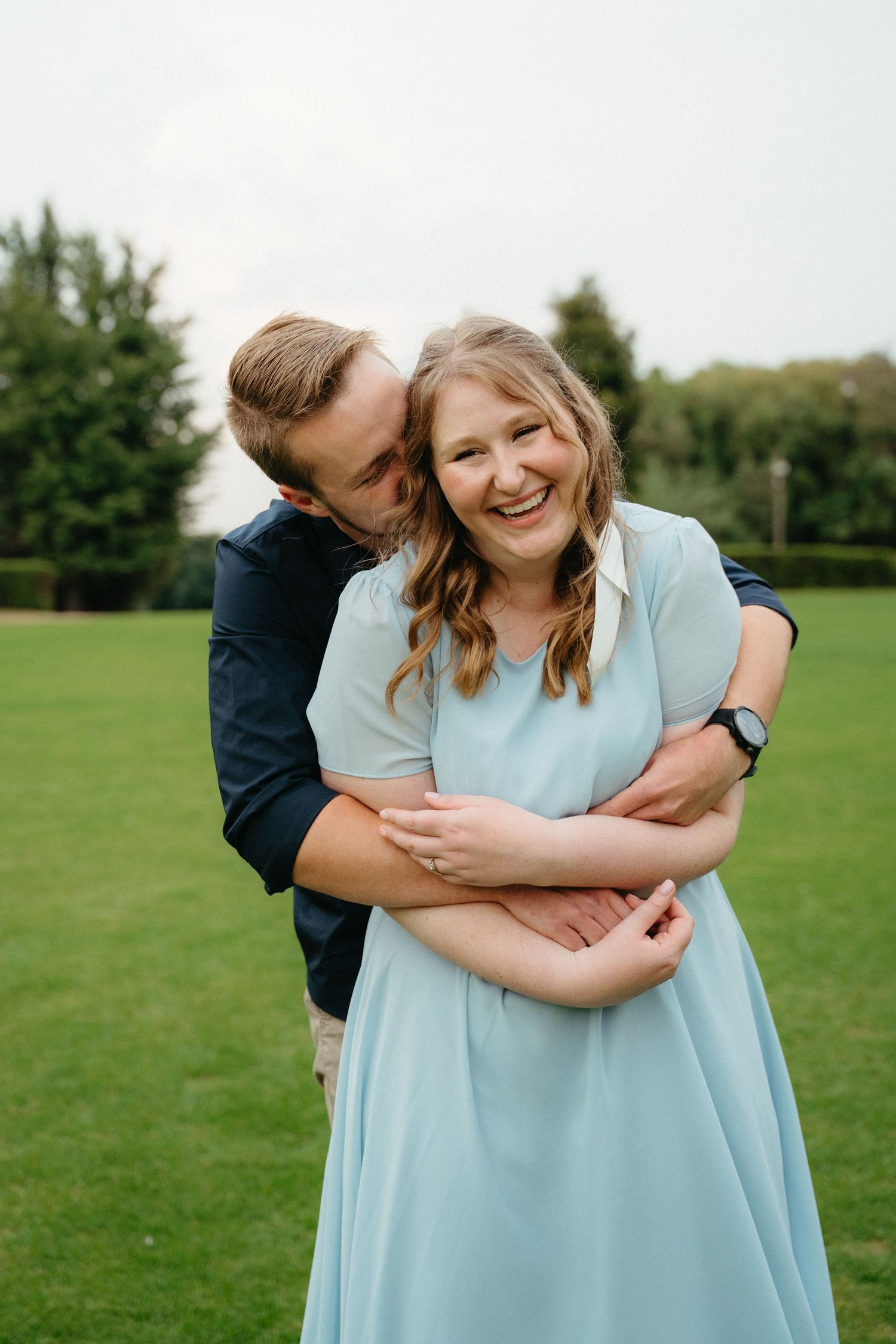 A couple embraces in a romantic moment on a grassy field while wearing light blue and dark clothing.