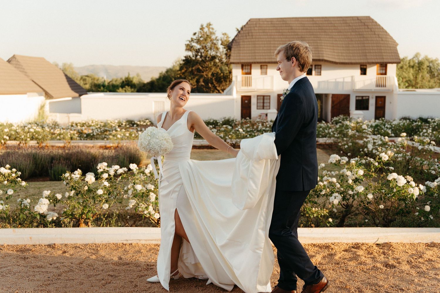 A couple in wedding attire walks together through a garden setting at sunset.