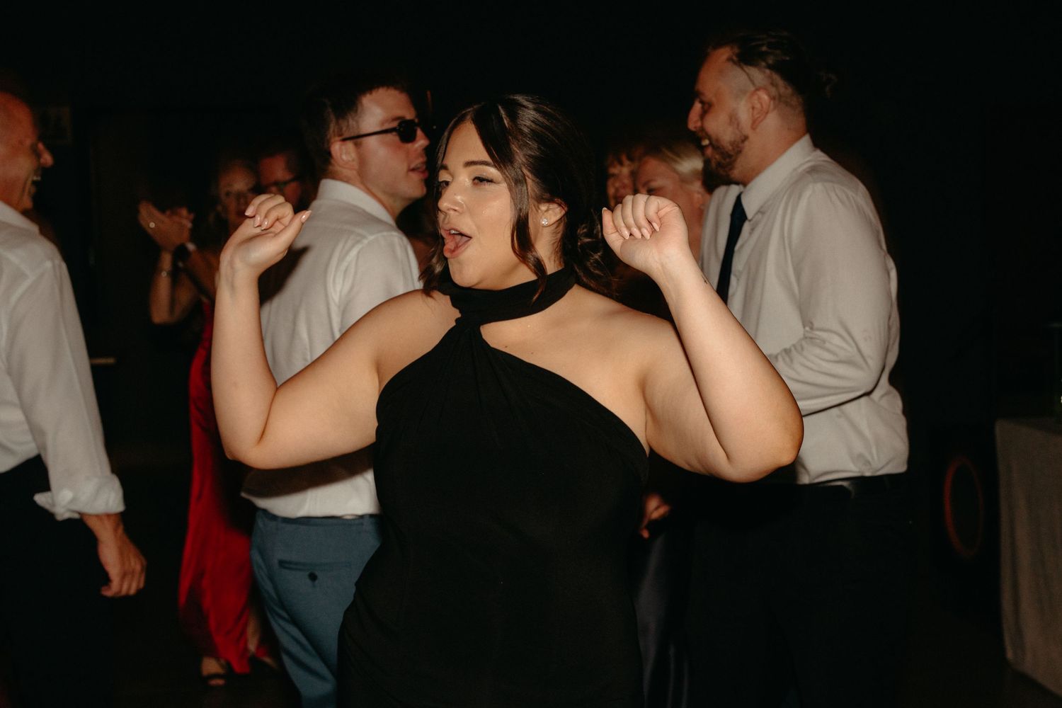 A person in a black dress dances enthusiastically with raised arms at a dark party venue.