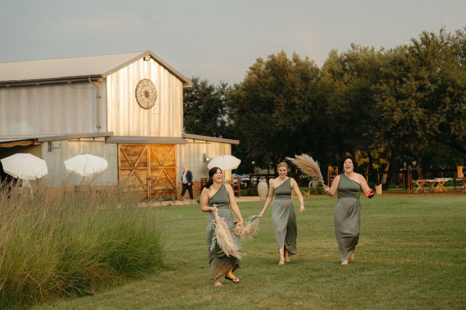 Wedding party in sage green dresses dance playfully across a rustic farm field at sunset.