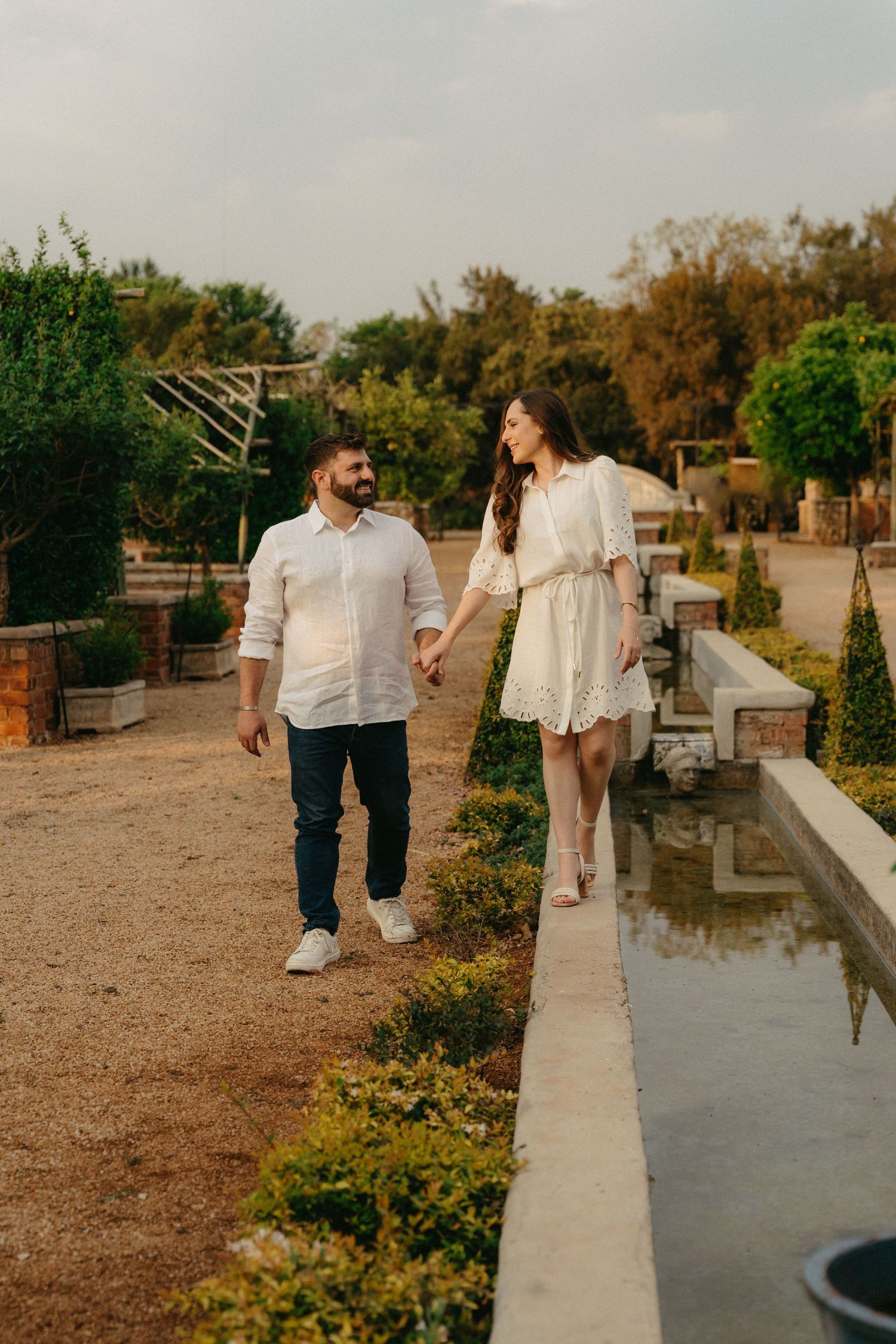 A couple walks along a concrete path in a garden setting during golden hour.