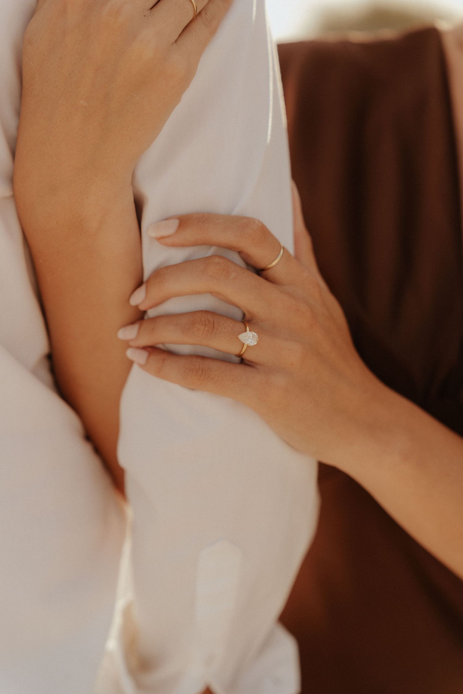 Close-up of a woman's hand wearing a gold solitaire diamond engagement ring, embracing someone in white and brown fabric.