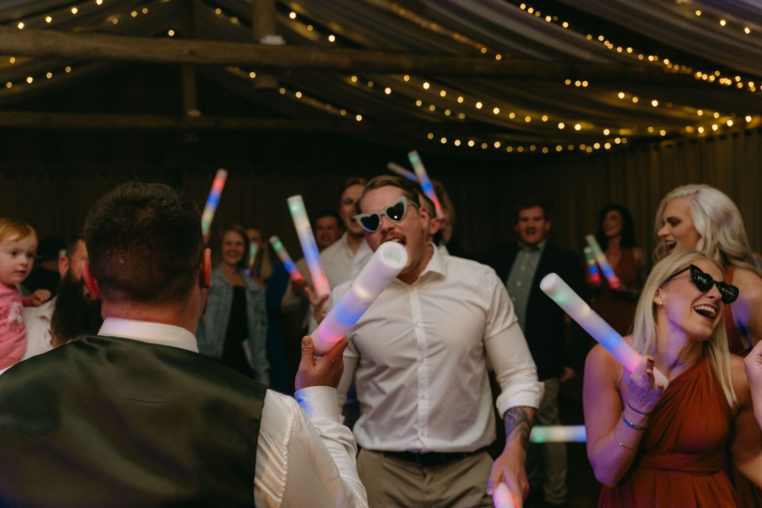 Wedding party guests dance and celebrate with colorful LED glow sticks under string lights at an evening reception.
