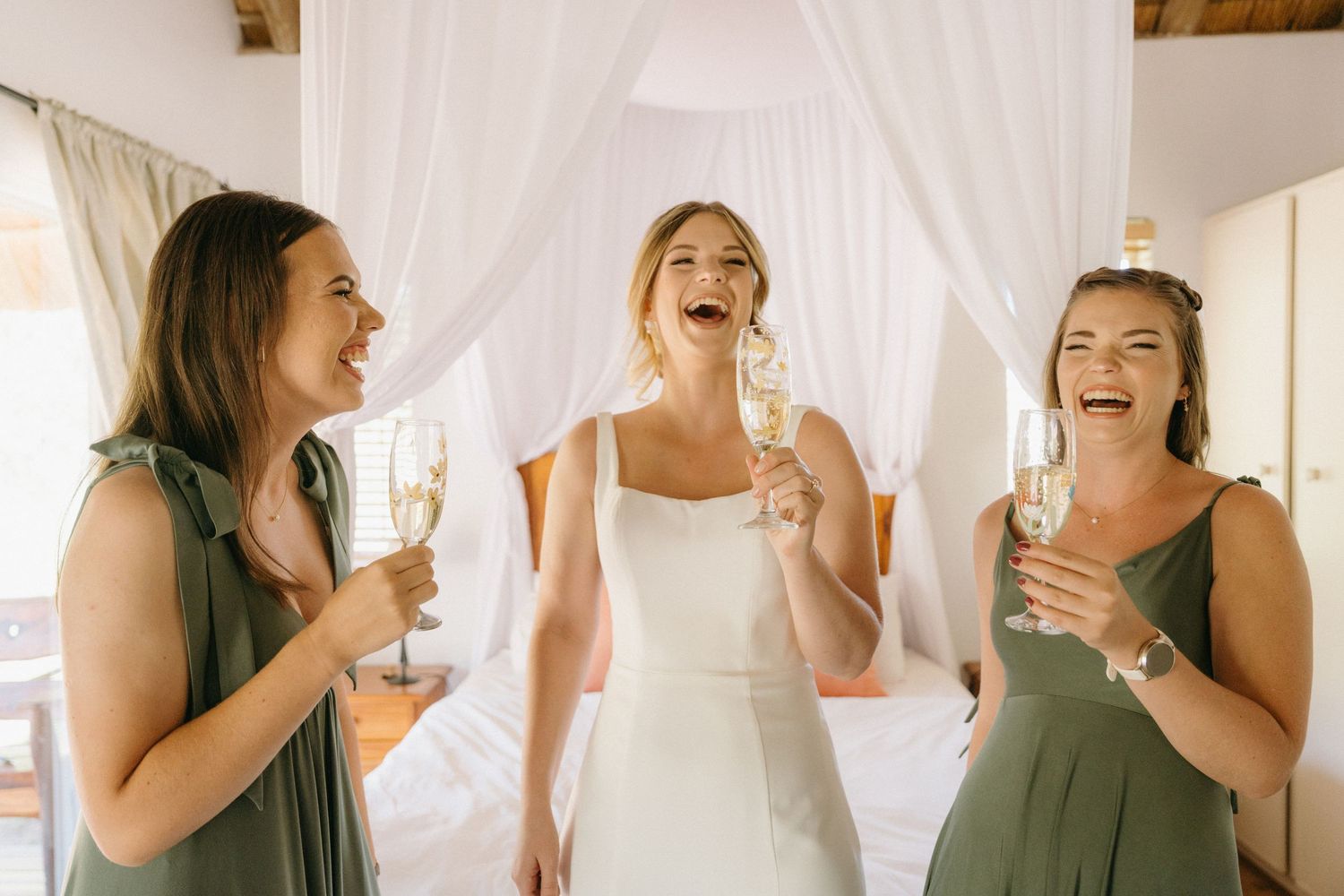 Three women in wedding attire sharing champagne and laughter in a bright room.