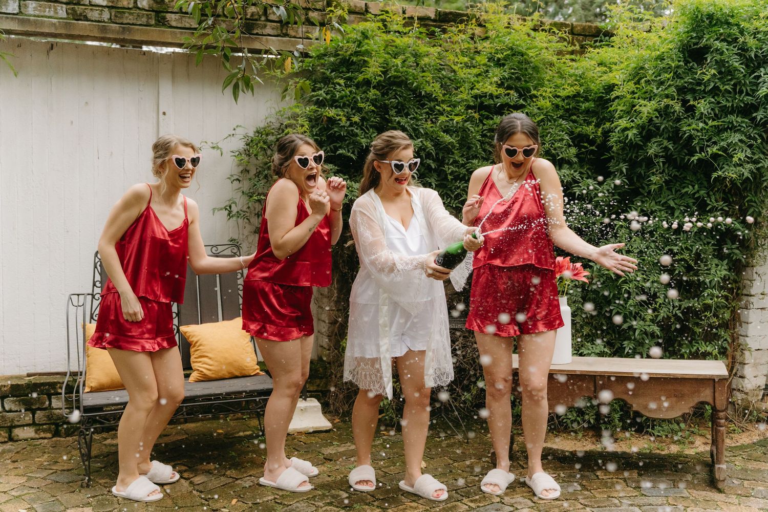 Bride in white robe laughing with bridesmaids wearing matching red robes while getting ready.