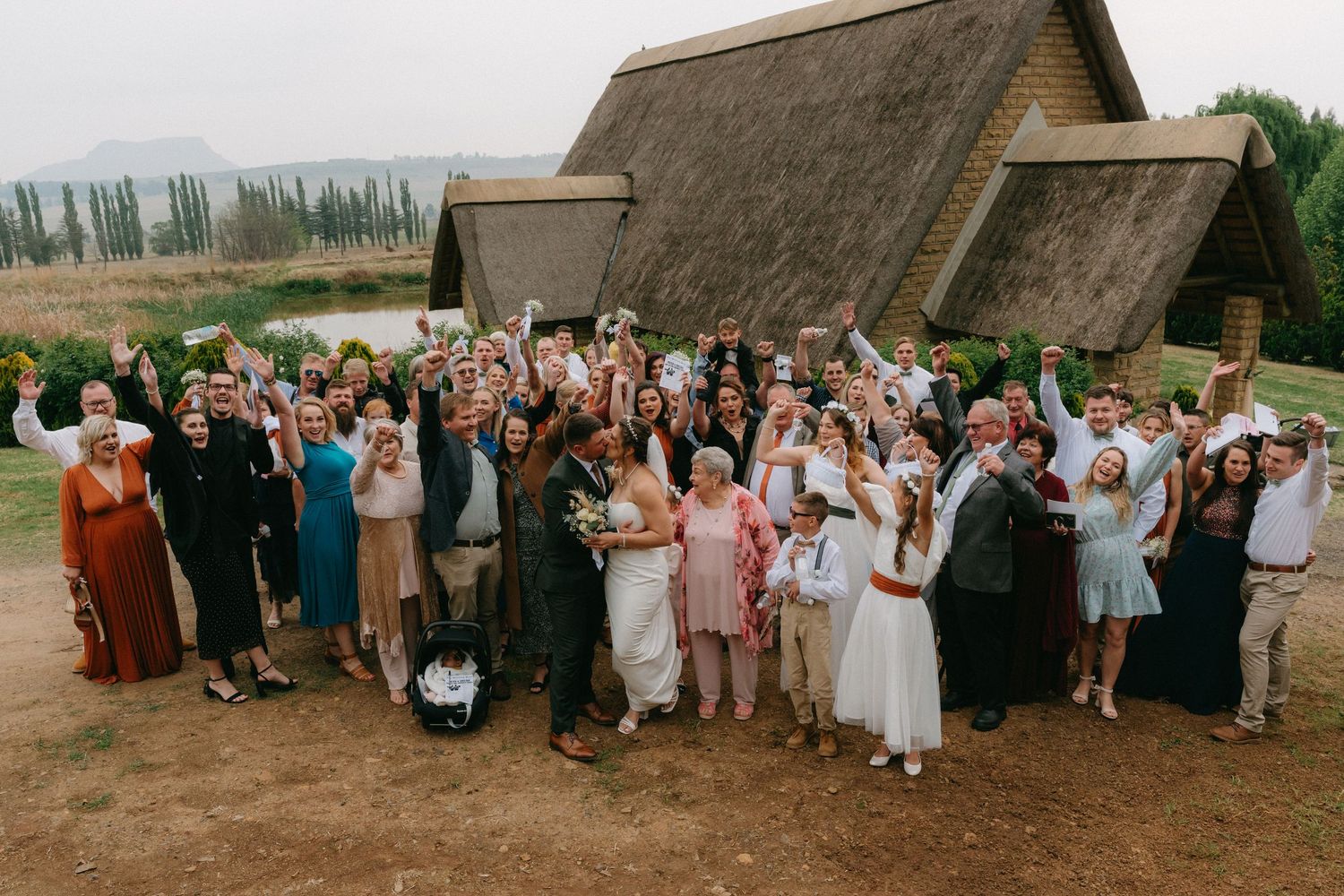 Large group of wedding guests gathered outside rustic barn venue with thatched roof in countryside setting.