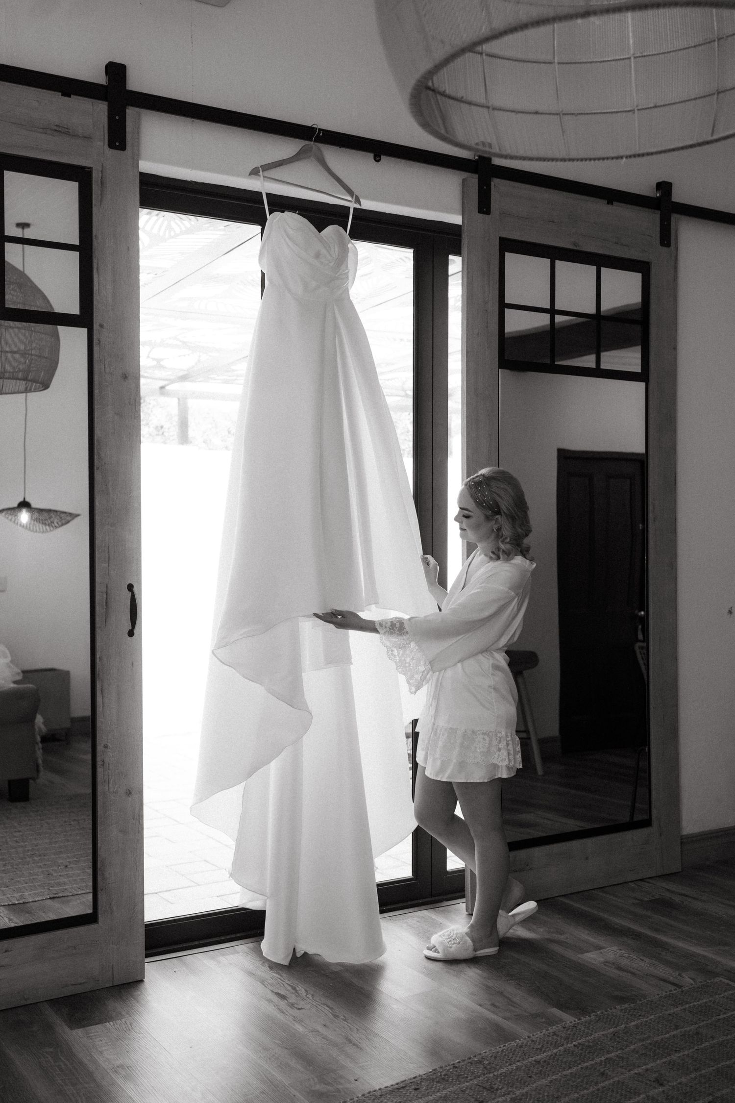 A white wedding dress hangs elegantly by a sliding glass door as someone adjusts it in black and white.
