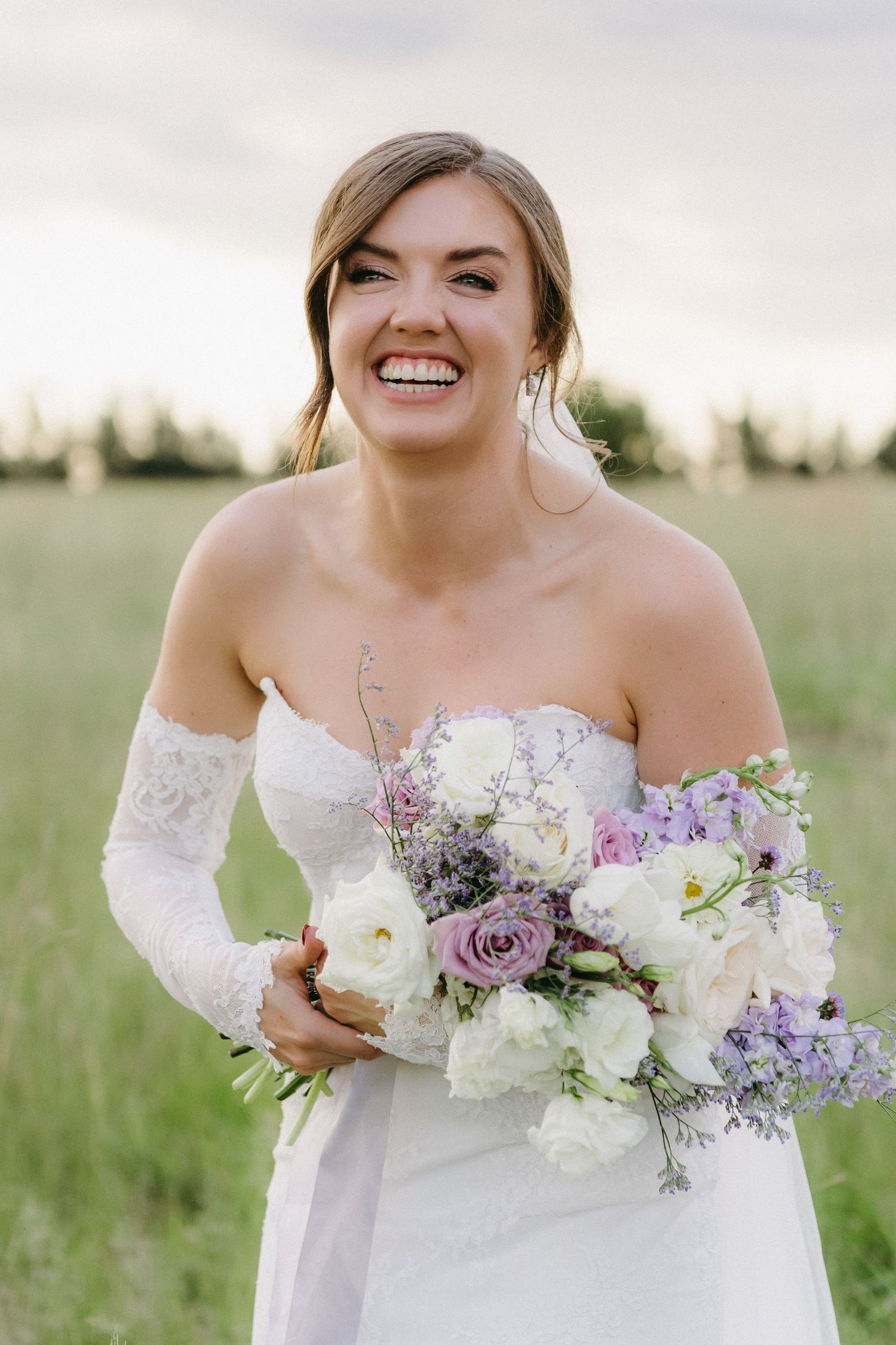 A radiant bride holding a purple and white bouquet smiles joyfully in a sunlit meadow.