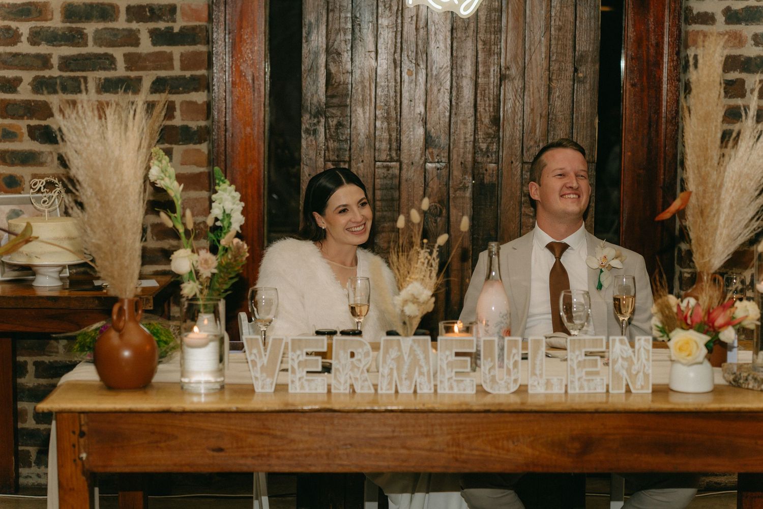 Wedding reception guests seated at long wooden tables decorated with floral centerpieces in a rustic brick venue.
