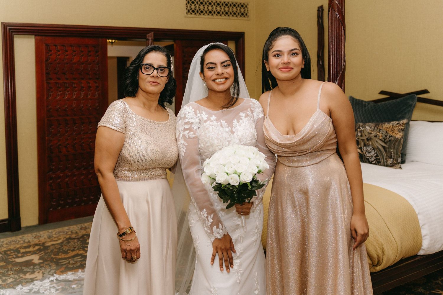 Three women in champagne-colored dresses pose together at a wedding, with the bride in white holding a bouquet of flowers.