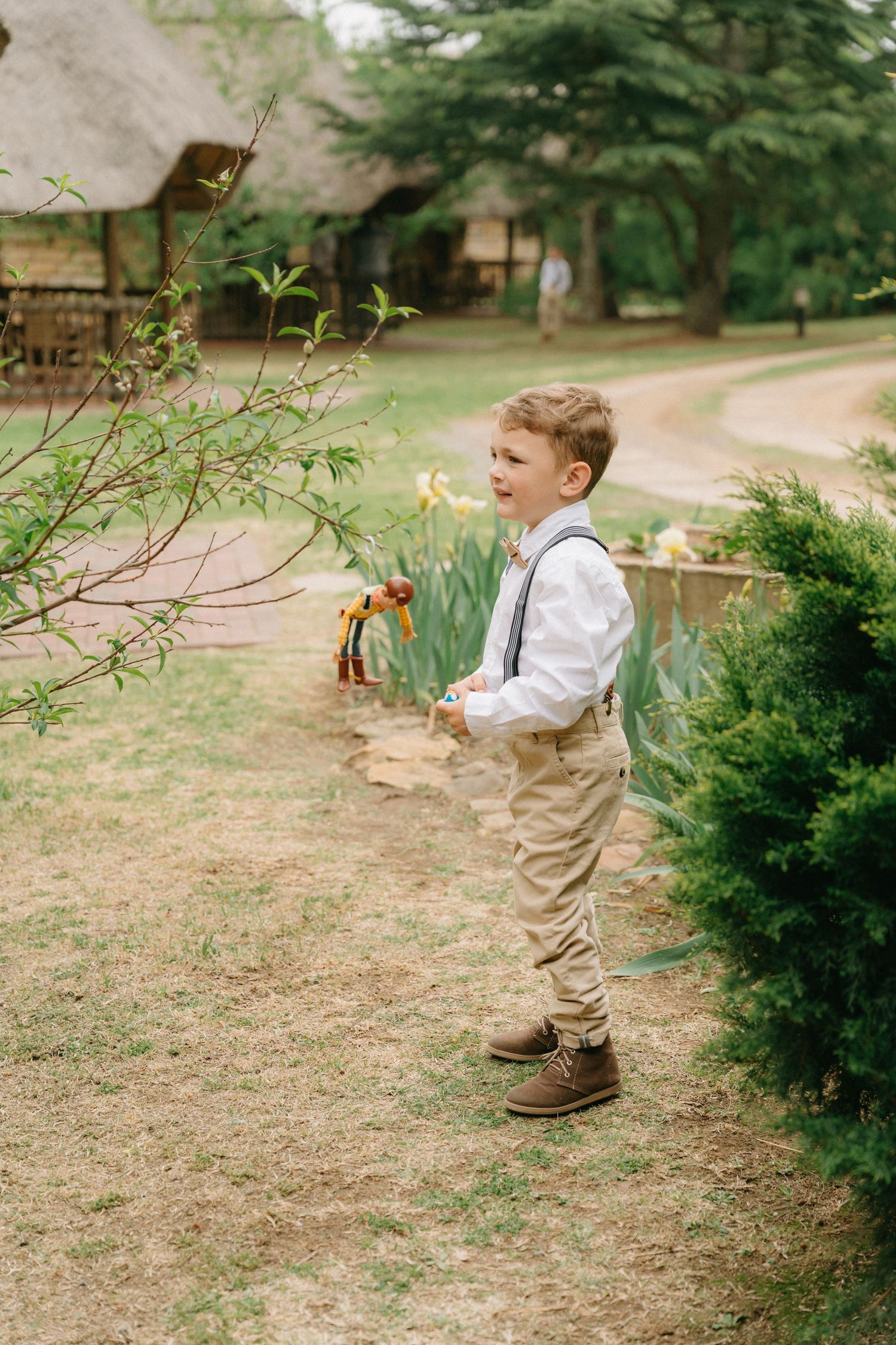 A young child in suspenders and dress clothes plays outdoors near bushes and flowers in a garden setting.