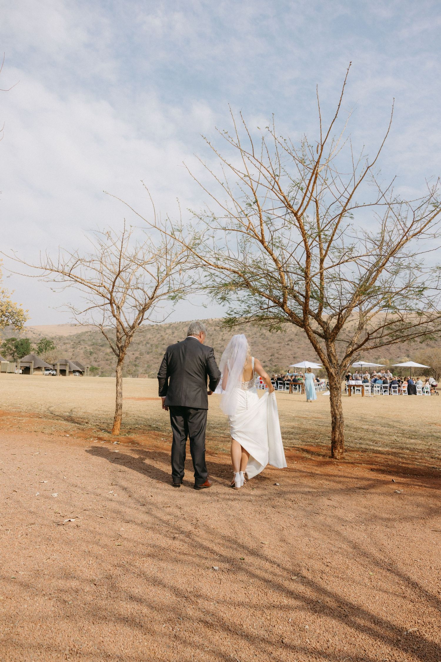 A couple walking between bare trees on a desert path with guests visible in the background.