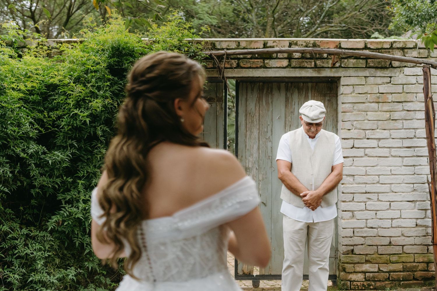 Emotional first look between bride in off-shoulder gown and groom in white against brick wall backdrop.