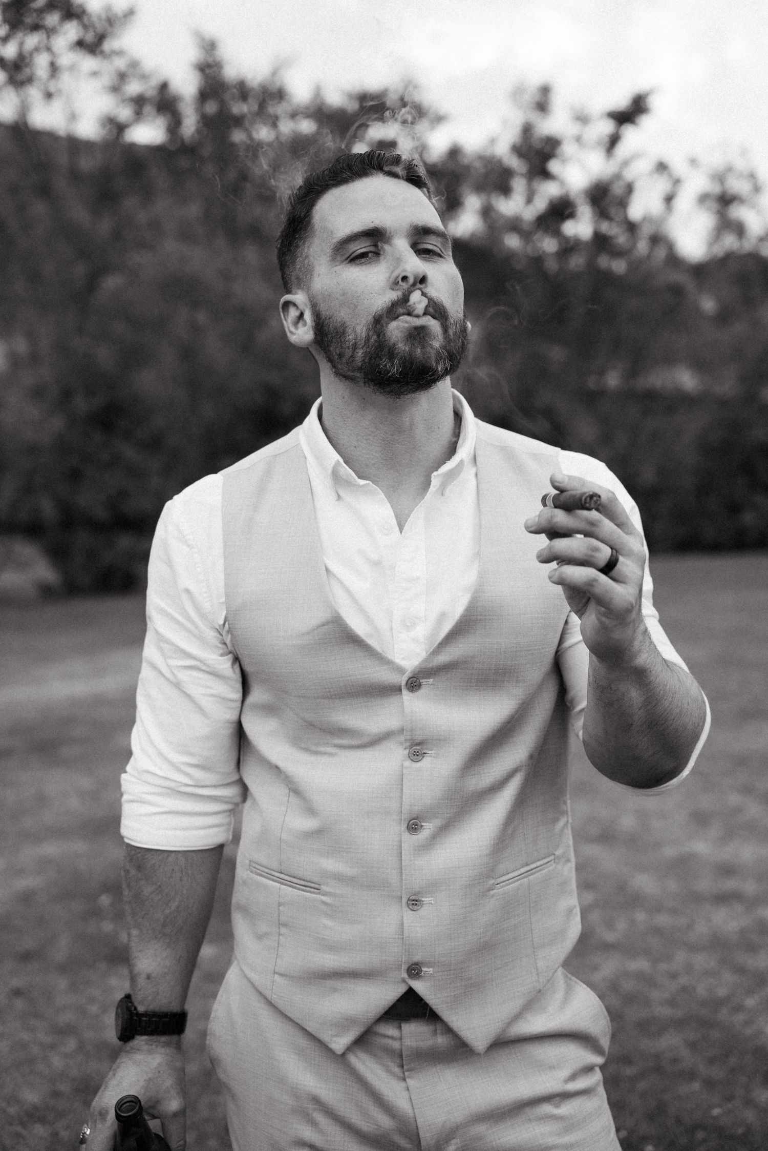 A groom in a light colored vest poses expressively outdoors.