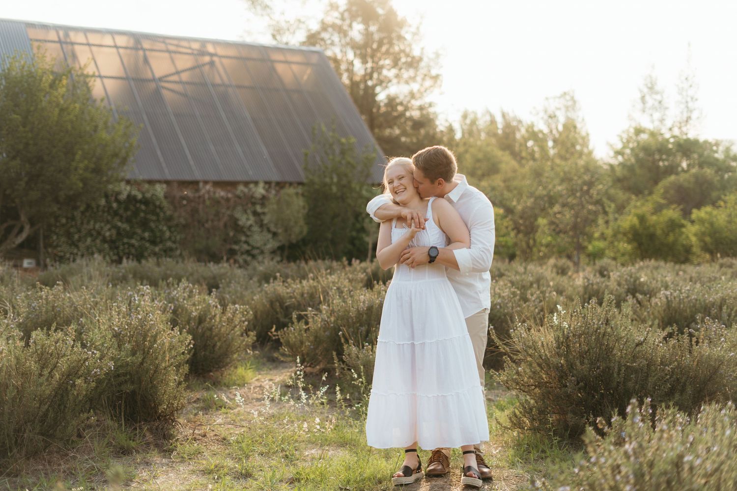 A couple embraces in a white sundress and casual attire during golden hour on their rustic outdoor photoshoot.