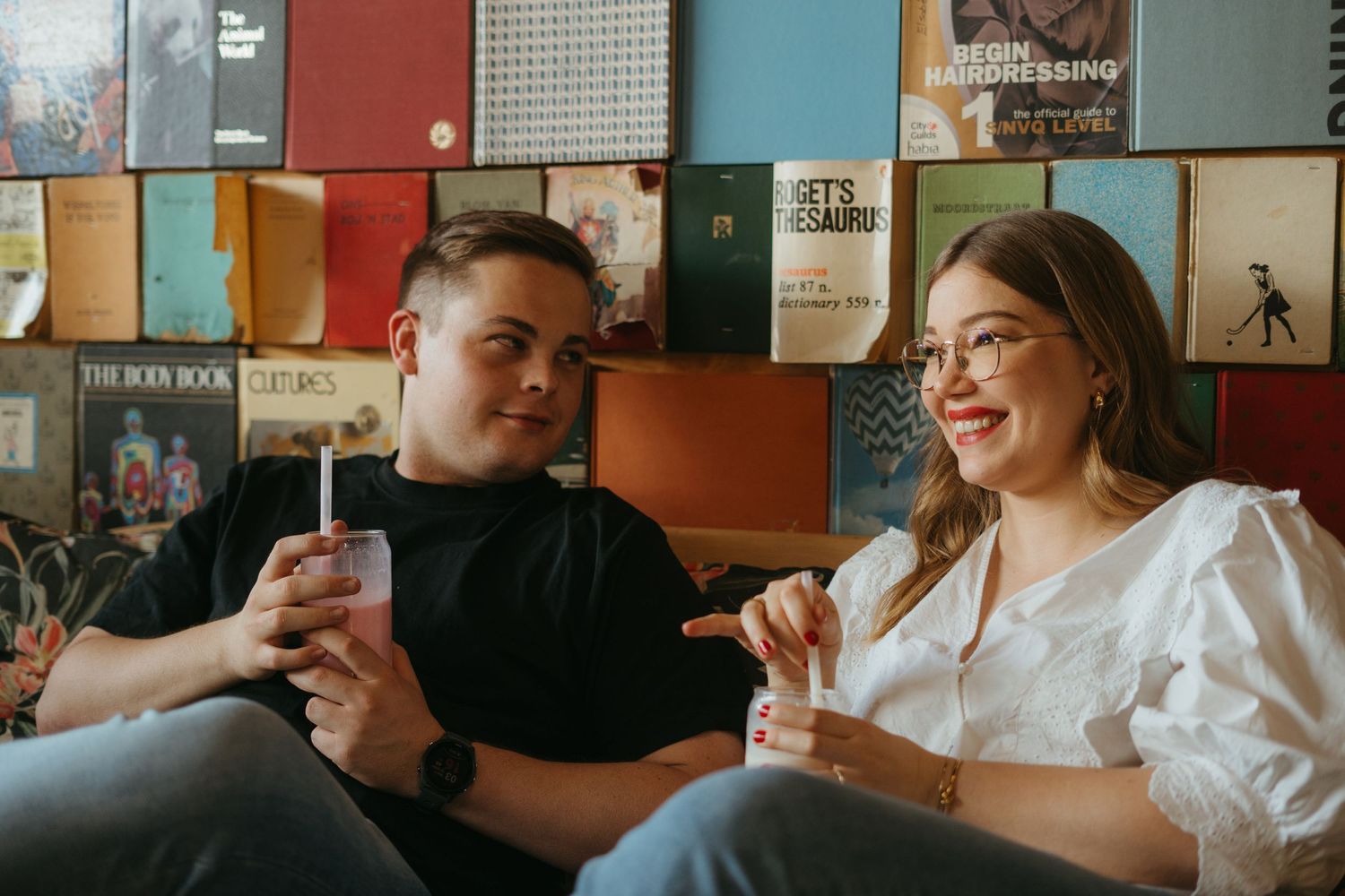 Two friends relax on a couch while enjoying drinks together against a wall decorated with colorful record albums.