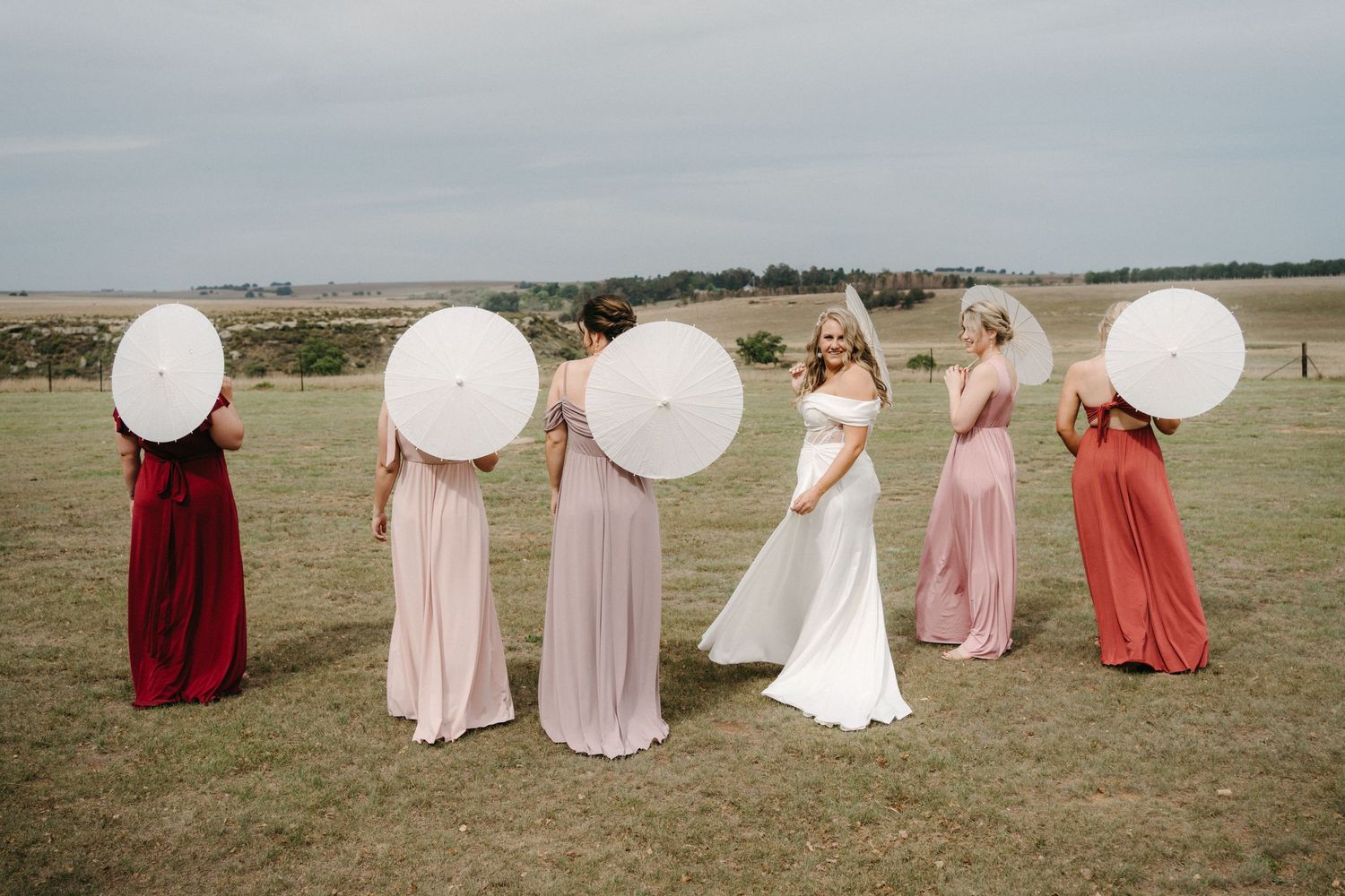 Wedding party members in burgundy and pink dresses pose with white parasols on a grassy field.
