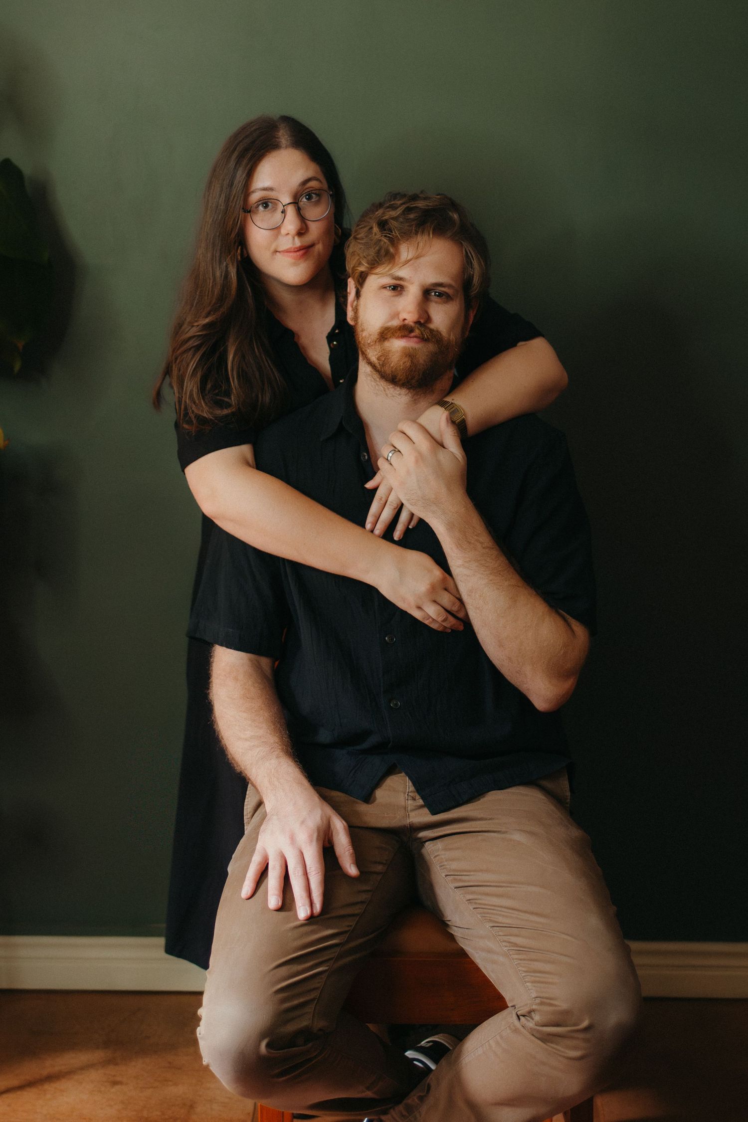 A  wedding photographer and videographer in black outfits share a warm embrace against a dark green wall.