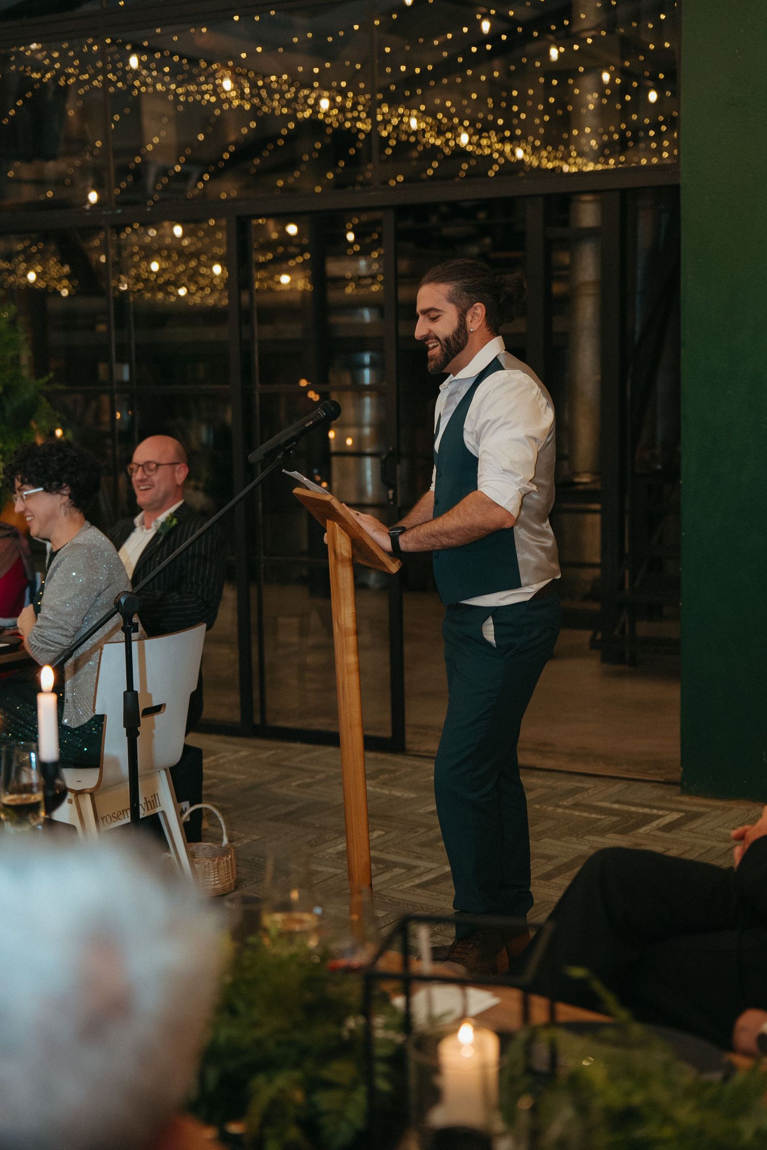 Smiling man giving a speech at a podium during an elegant evening event with fairy lights.