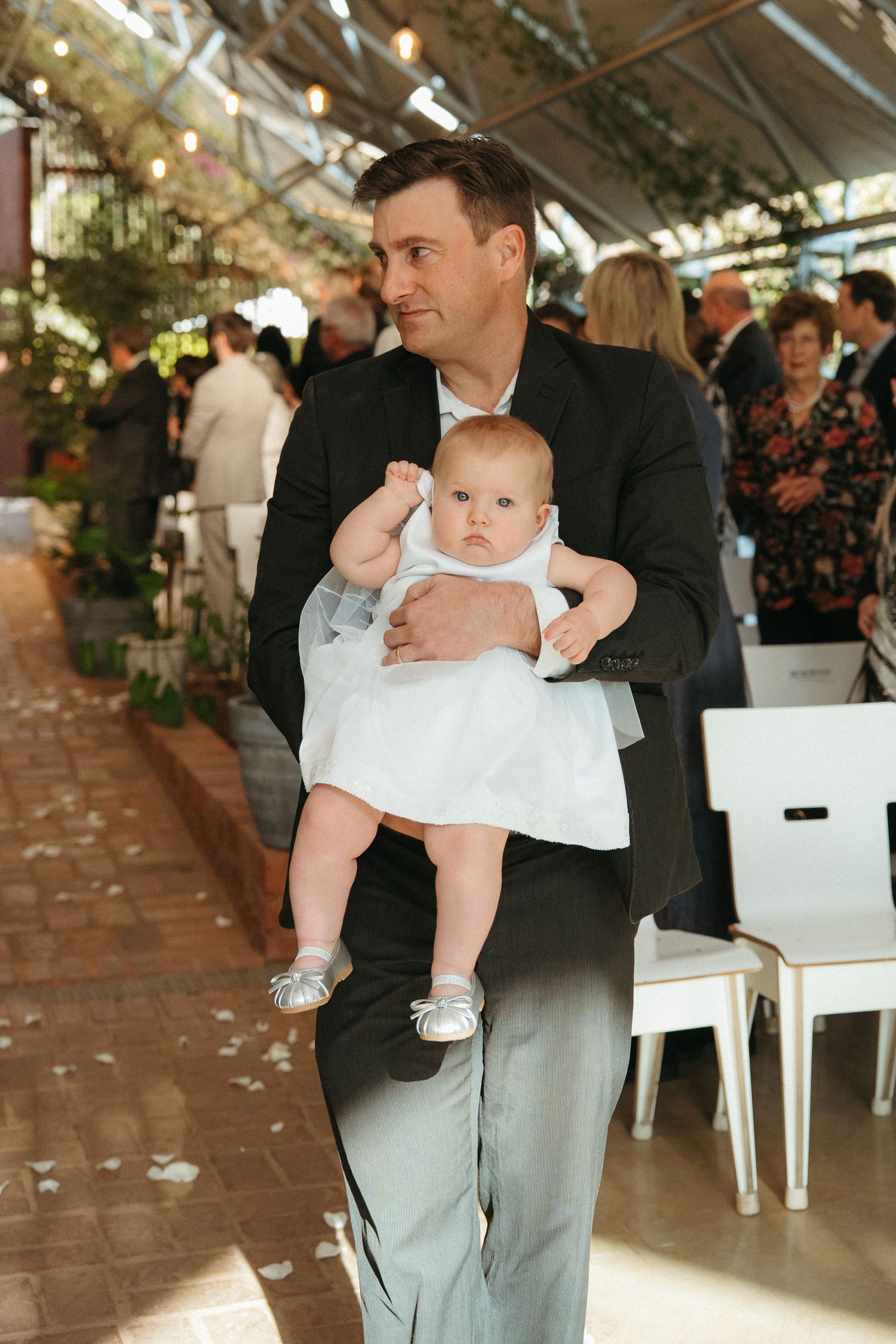 Man in black suit holding baby girl in white dress at an elegant indoor garden wedding ceremony.