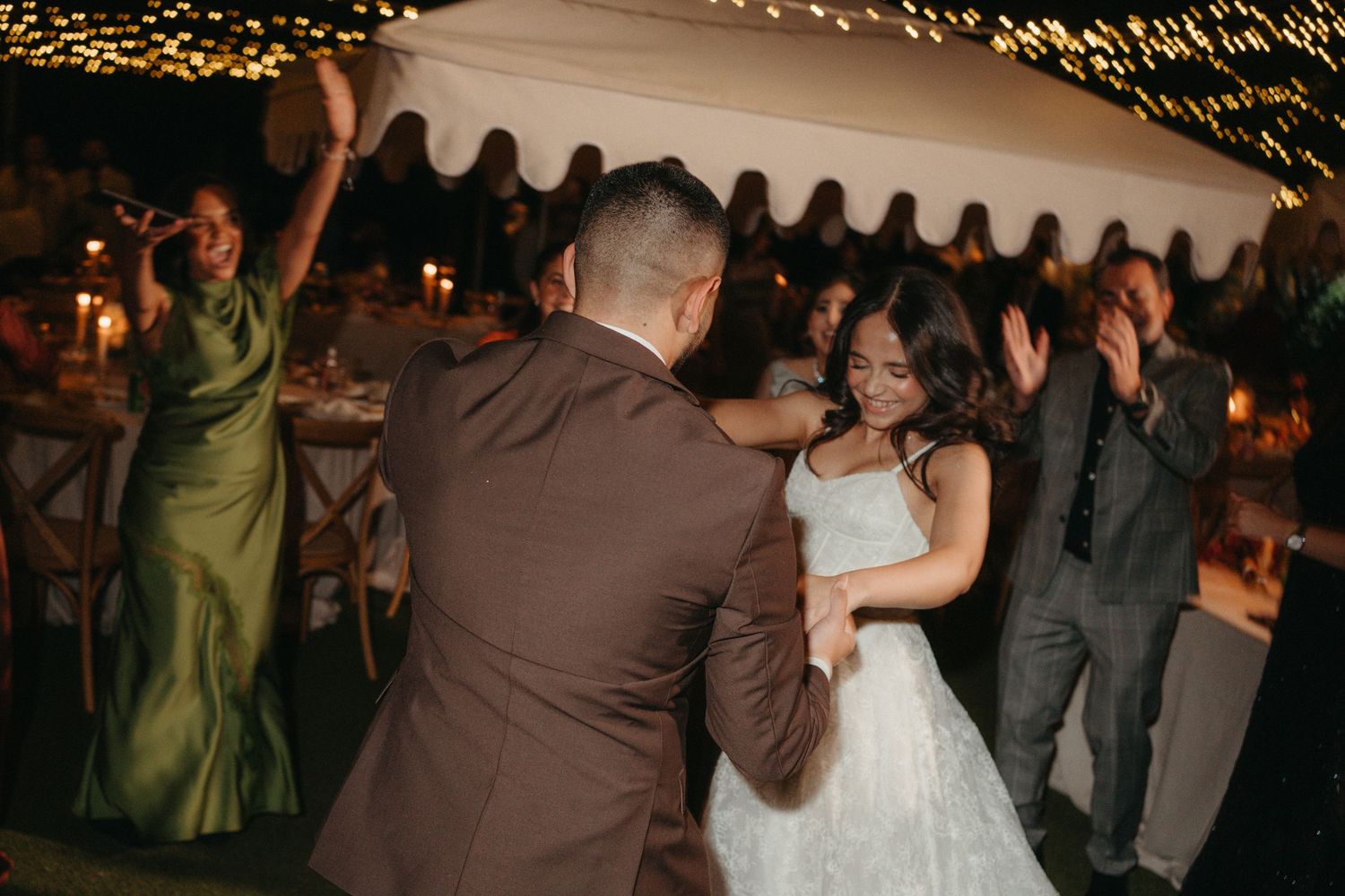 Bride and groom share first dance at elegant outdoor wedding reception with candles and guests celebrating.
