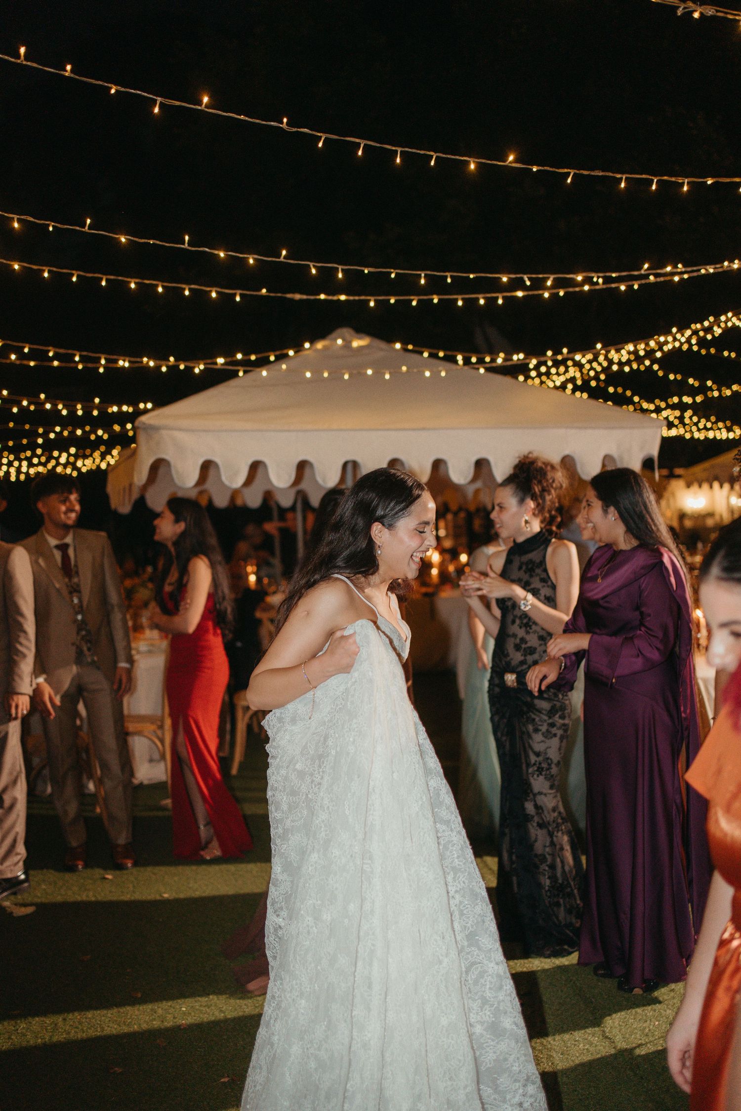 Bride in white lace gown dancing joyfully at outdoor wedding reception with string lights overhead.
