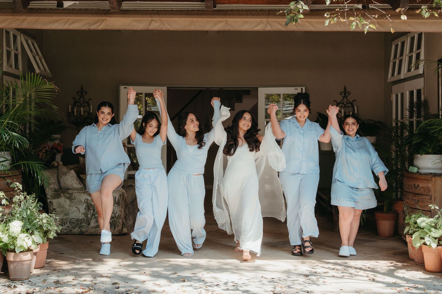 Bride in white gown celebrates with bridesmaids in matching light blue pajamas, arms raised joyfully outdoors.
