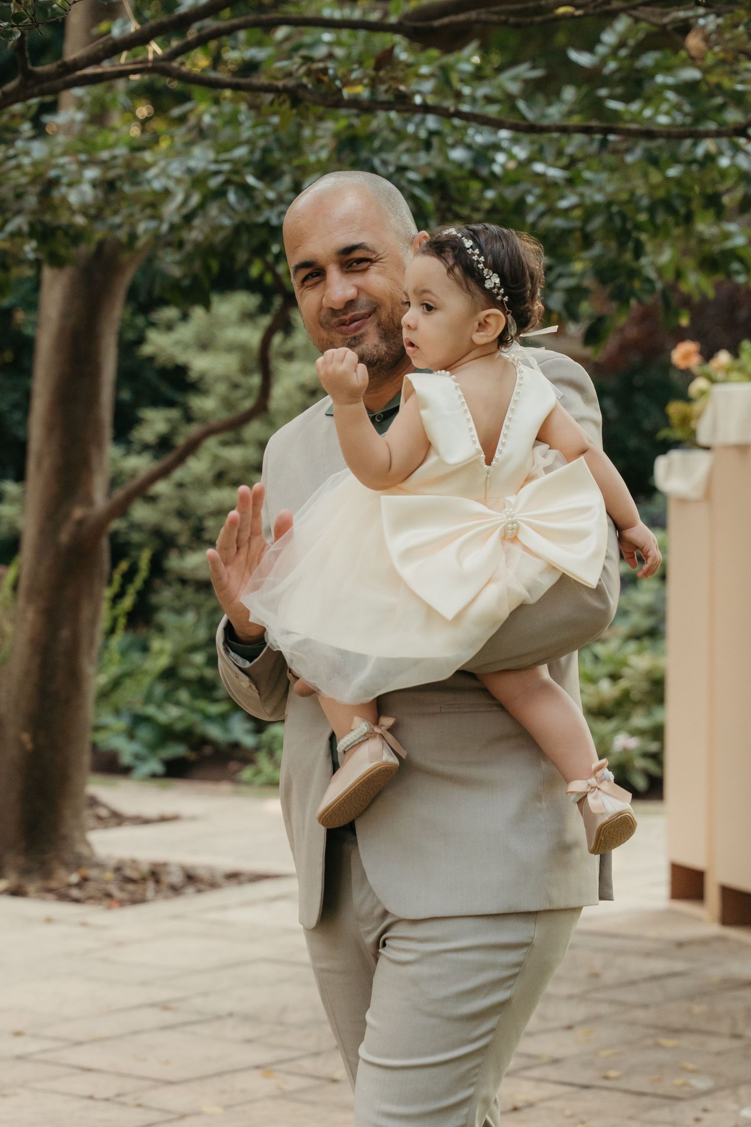 Bald man in beige suit holding a baby girl in white dress with bow outdoors in garden setting.
