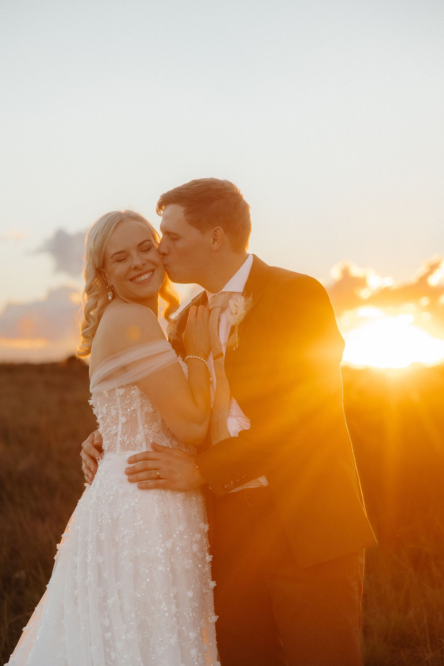 Groom kissing bride on cheek at golden sunset, bride smiling in white floral gown, romantic outdoor wedding portrait.
