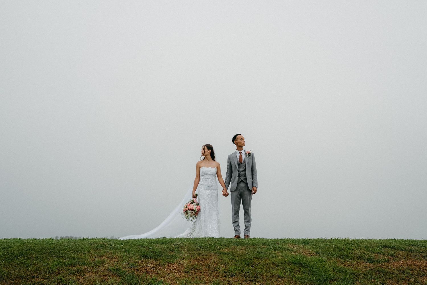 A bride and groom hold hands on a grassy hilltop against a misty sky on their wedding day.