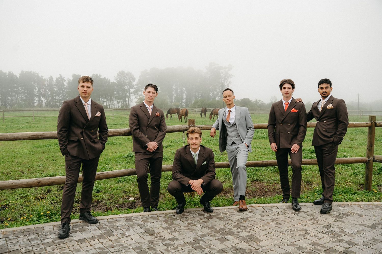 Six groomsmen and a groom in brown and gray suits pose by a rustic wooden fence on a foggy Tafika Polo Farm with horses grazing behind them.