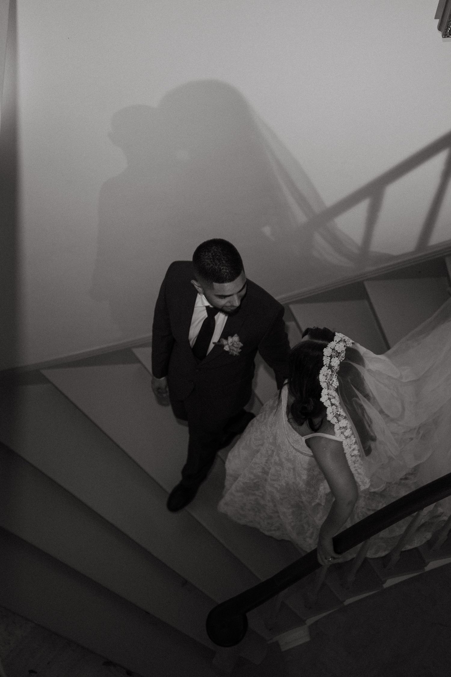 Black and white aerial view of bride and groom holding hands on staircase, shadows cast on wall behind them.