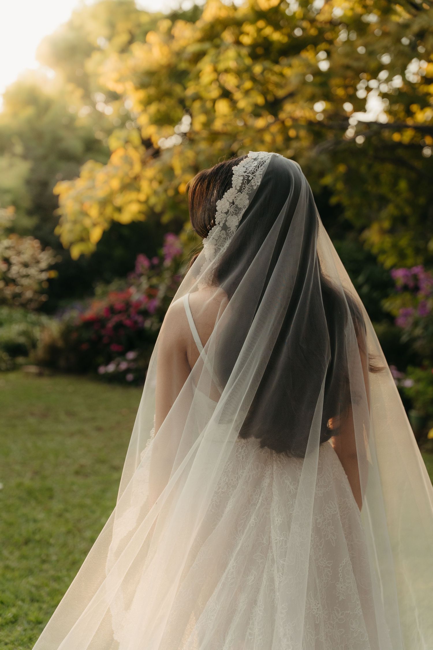Bride in white lace gown and flowing veil with floral headpiece stands in lush garden at golden hour.