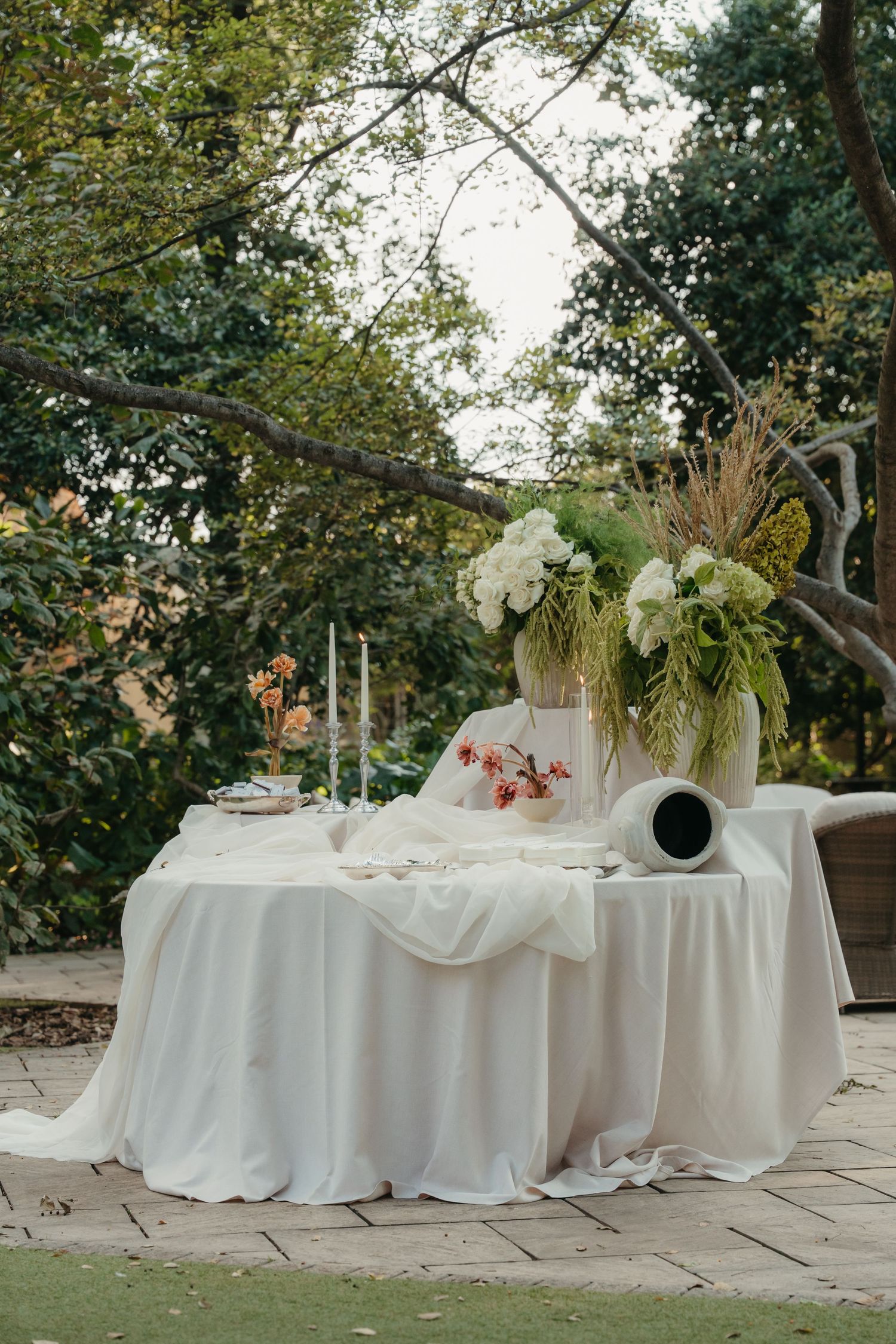 Elegant outdoor wedding table with white linen, candles, and floral arrangements in a lush garden setting.