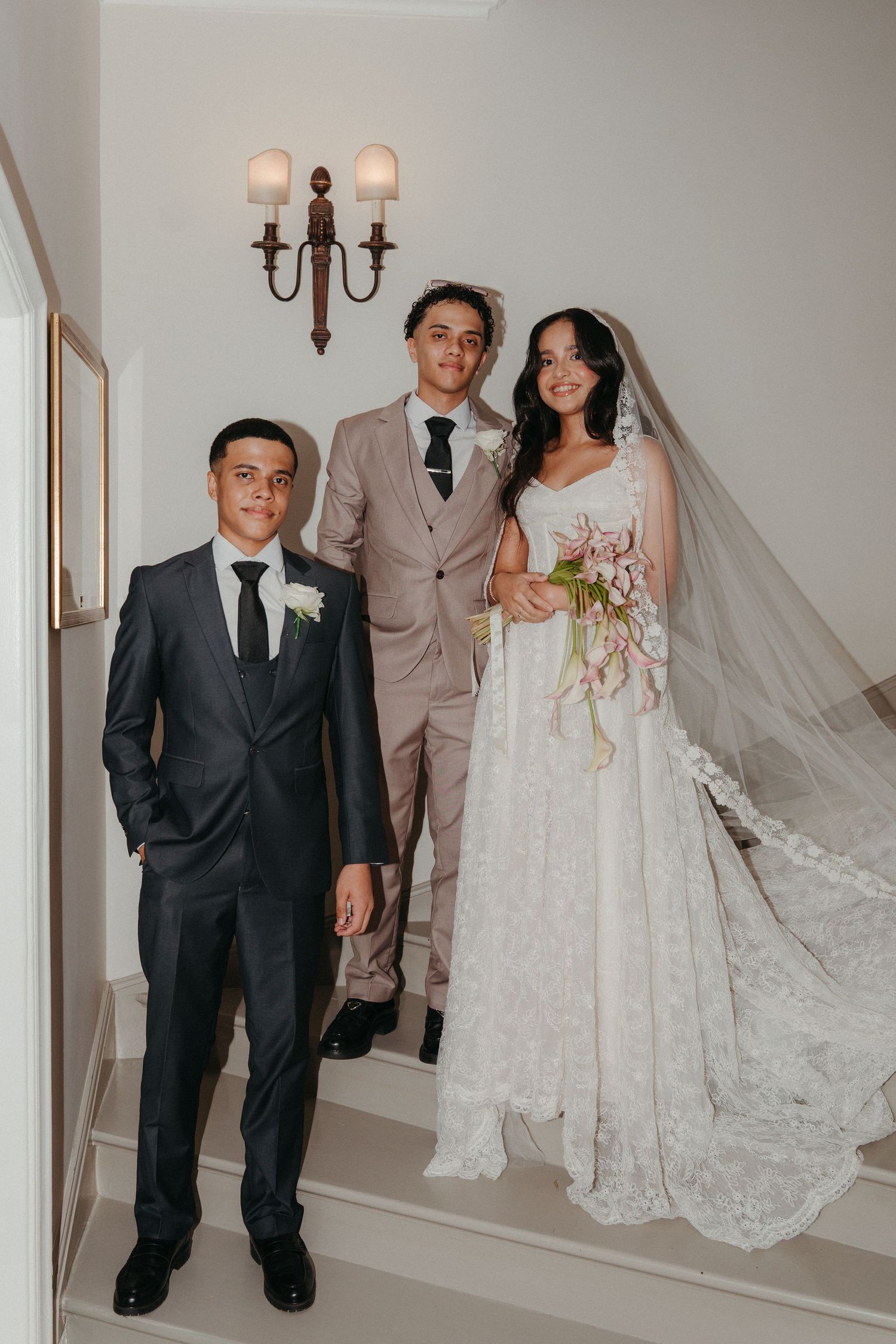 Bride in lace gown with veil holding bouquet poses with two groomsmen in suits on elegant white staircase.